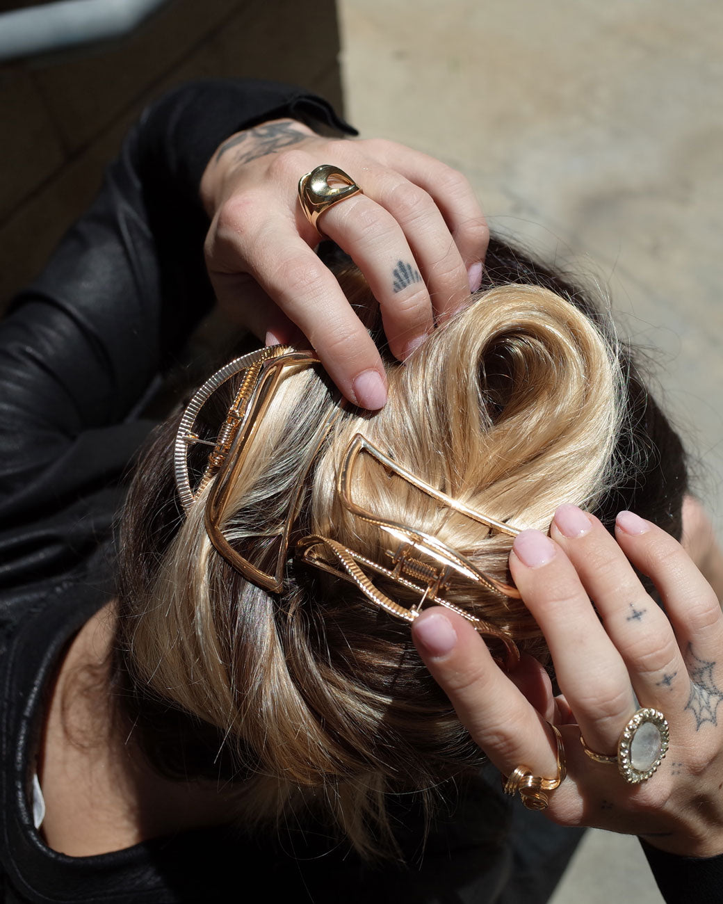 A woman's blond hair twisted up in two gold hair clips with spring loaded tops and teeth at the bases. Shown from the top with her hands holding her head.