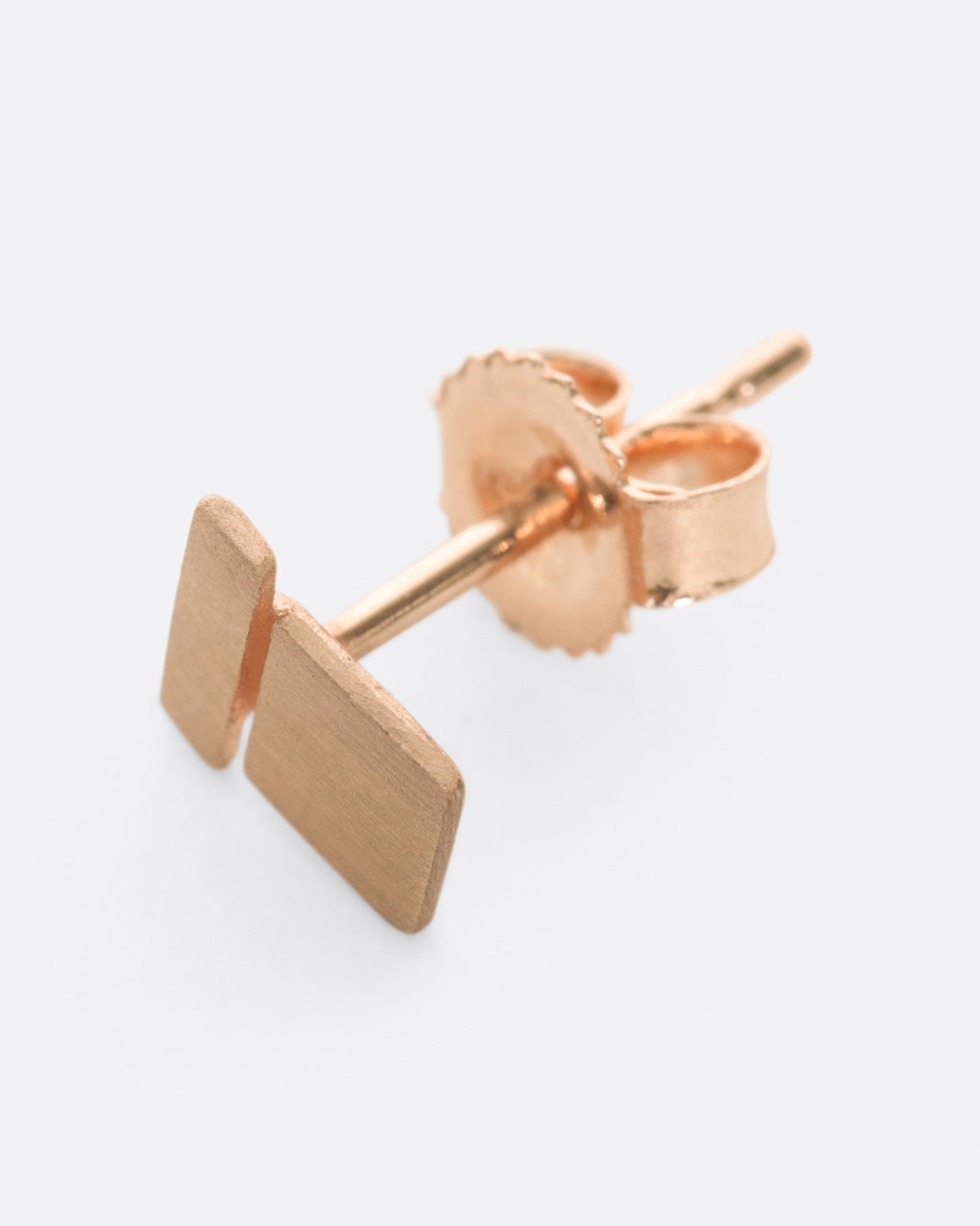 A matte rose gold stud earring with a bar stacked on a square. Shown on its side at an angle from the front.