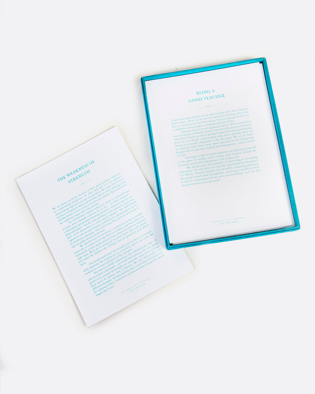 A turquoise box open, displaying two white cards with turquoise lettering. Shown lying flat at an angle from the top.