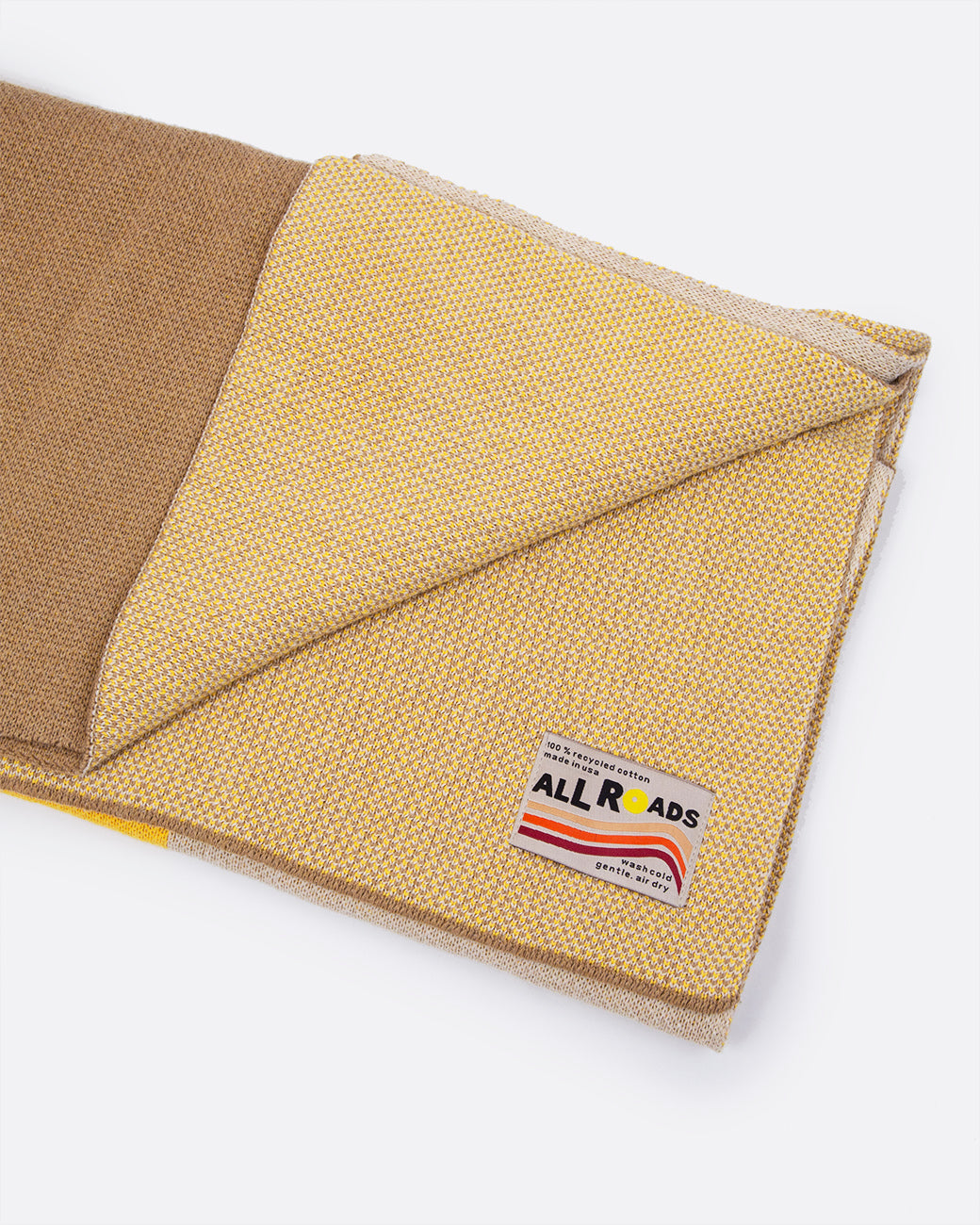 A yellow, tan, and brown blanket folded with a corner turned up to show the tag. The tag reads: 'ALL ROADS' with orange and red stripes at the bottom. Shown lying flat at an angle.
