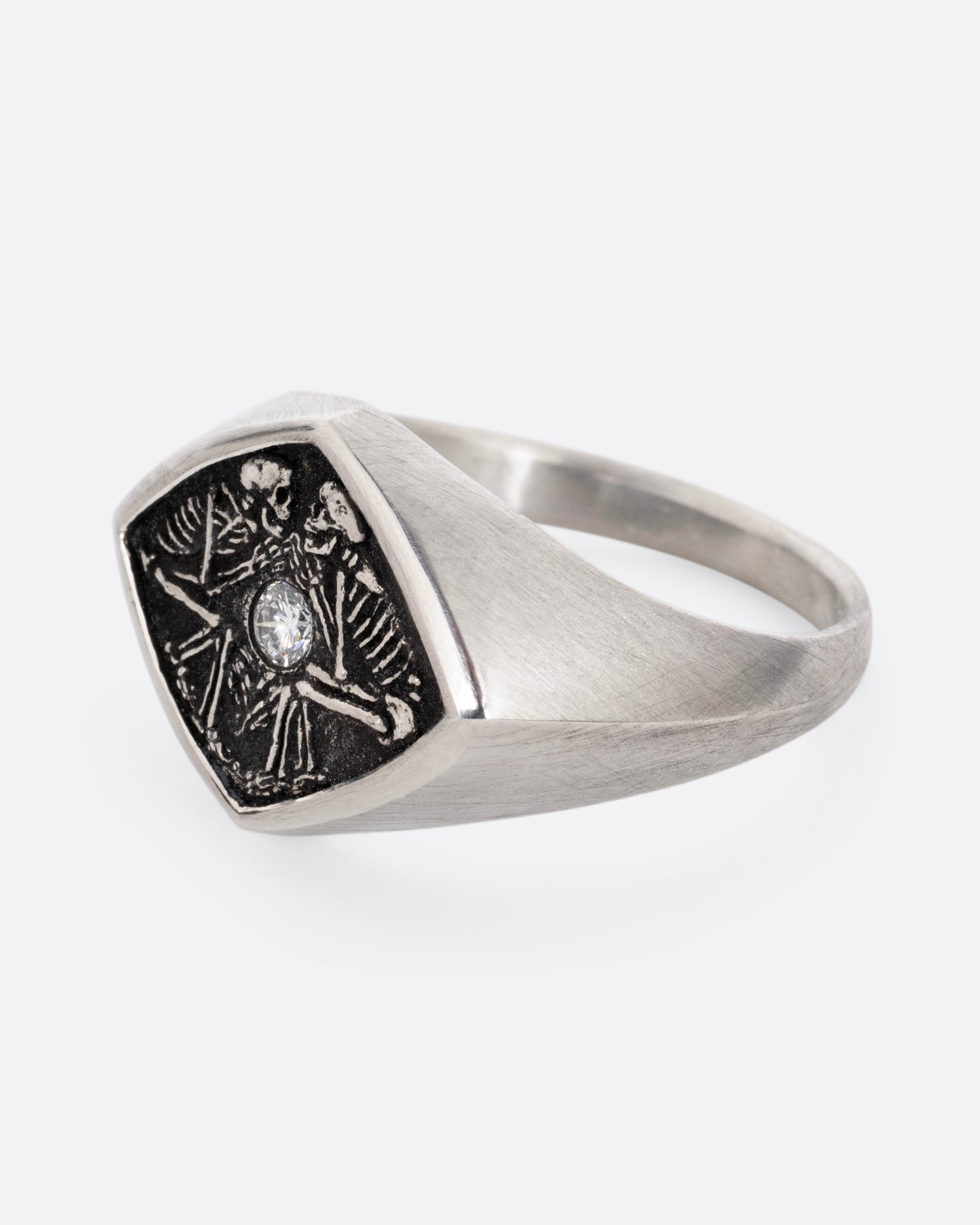 A silver signet ring with two skeleton figures in an oxidized backdrop with a white flush set diamond between the pair. Shown from the side.