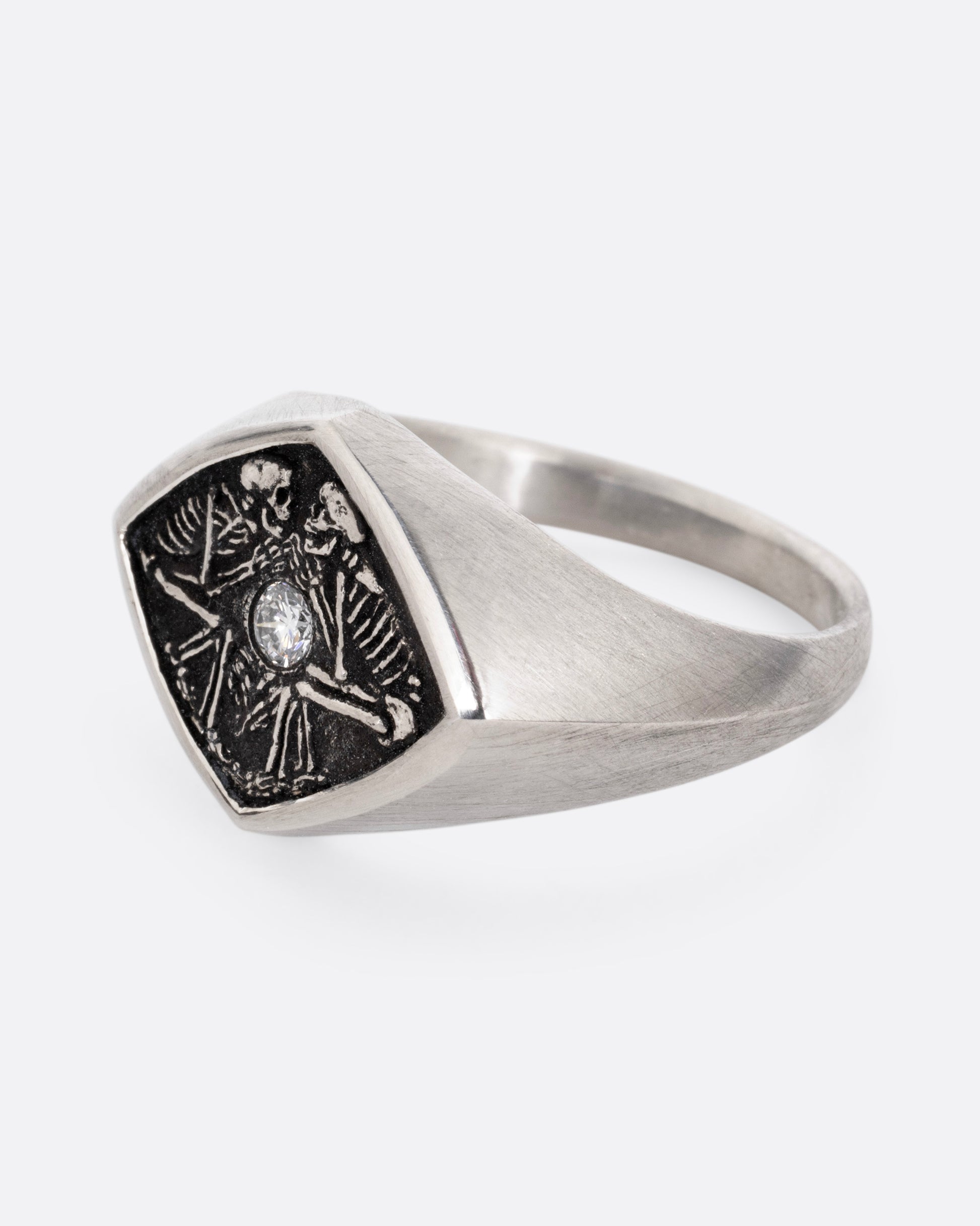 A silver signet ring with two skeleton figures in an oxidized backdrop with a white flush set diamond between the pair. Shown from the side.