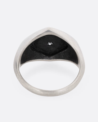 A silver signet ring with two skeleton figures in an oxidized backdrop with a white flush set diamond between the pair. Shown from the back.