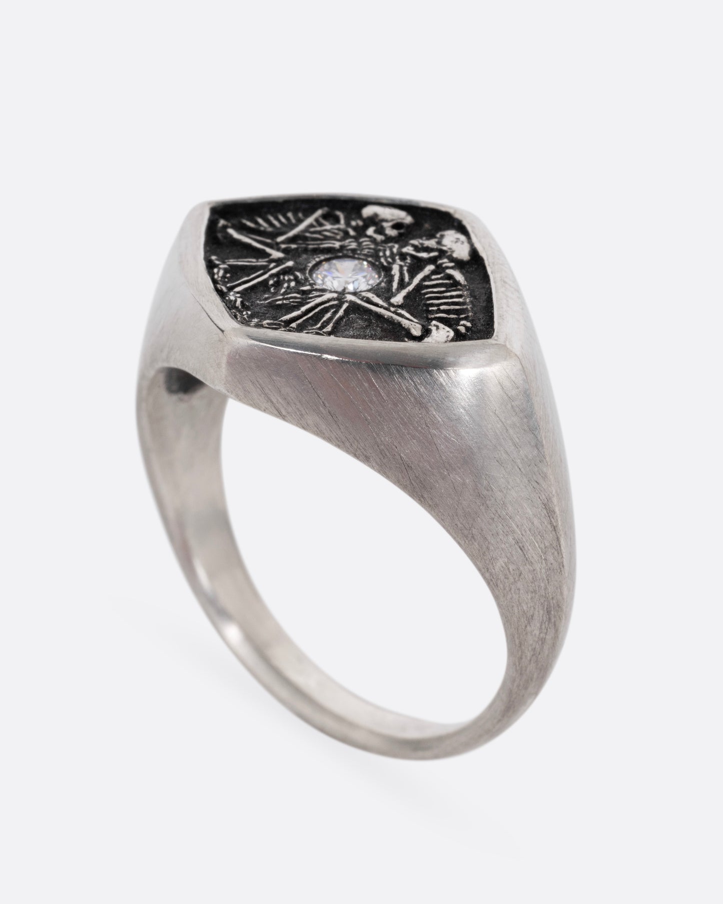 A silver signet ring with two skeleton figures in an oxidized backdrop with a white flush set diamond between the pair. Shown from the side standing up.