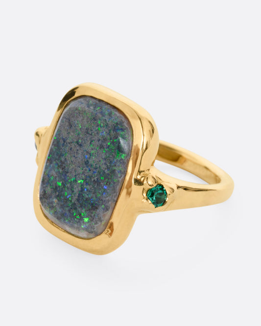 A yellow gold ring with a large rectangular boulder opal and emerald accents, shown from the side.