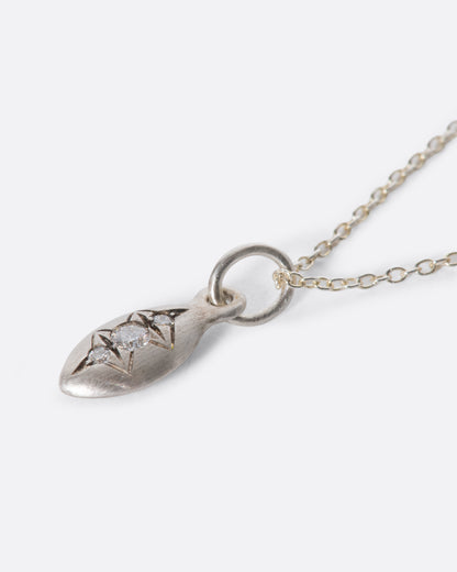 A silver cable chain necklace with a seed shaped pendant with three diamonds and oxidized etching. Shown lying flat at an angle from the side.