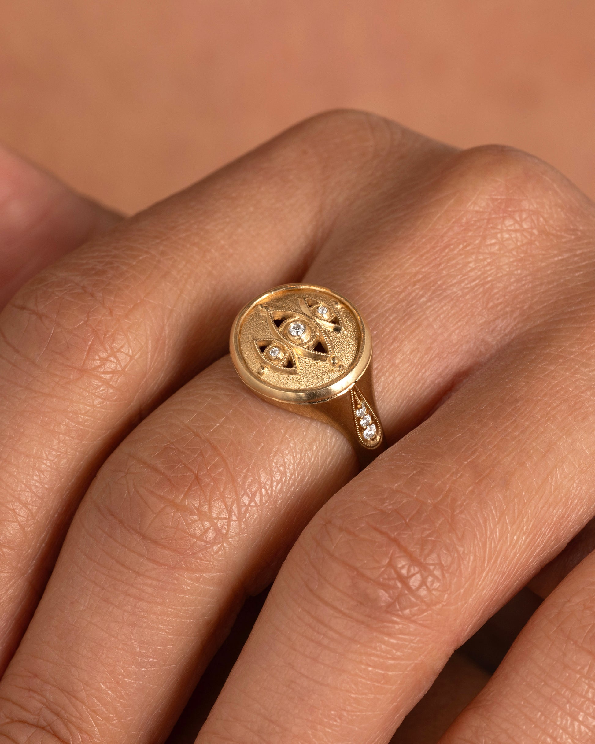 A yellow gold signet ring with three relief carved diamond eyes at the center. A trio of diamonds in teardrop shaped settings top each shoulder. Shown worn on a finger.