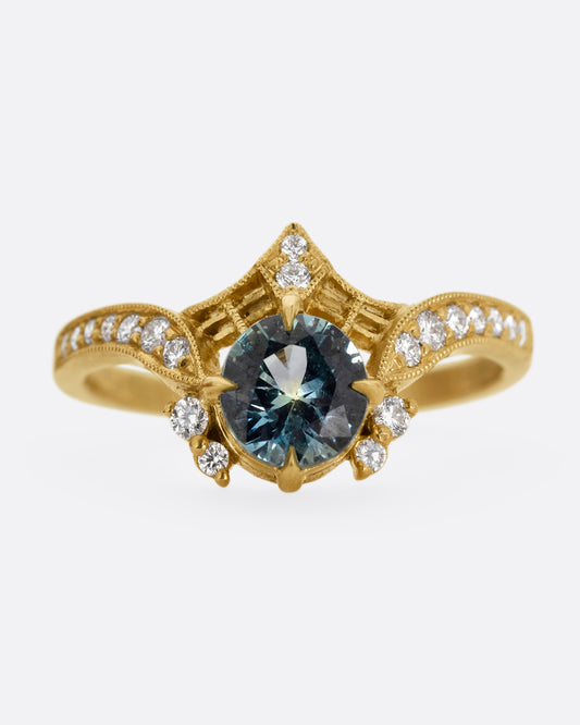 A yellow gold ring with a prong set teal sapphire, delicate cutouts, and white diamond accents. Shown from the front.