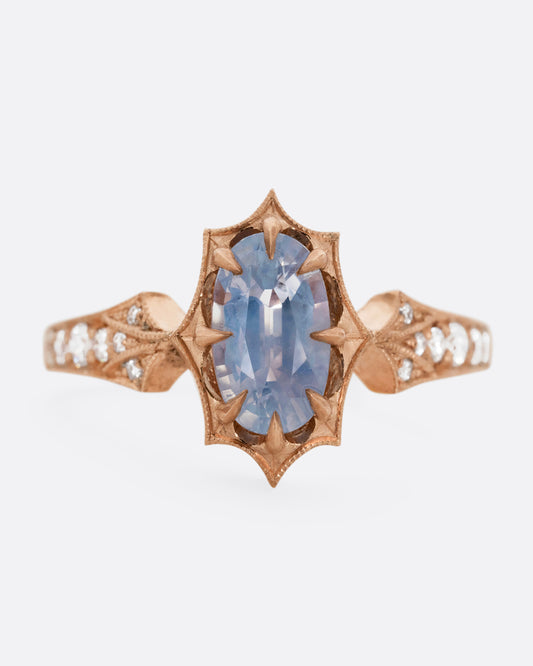 A rose gold ring with a prong set lavender sapphire at the center and diamond lined shoulders. Shown from the front.