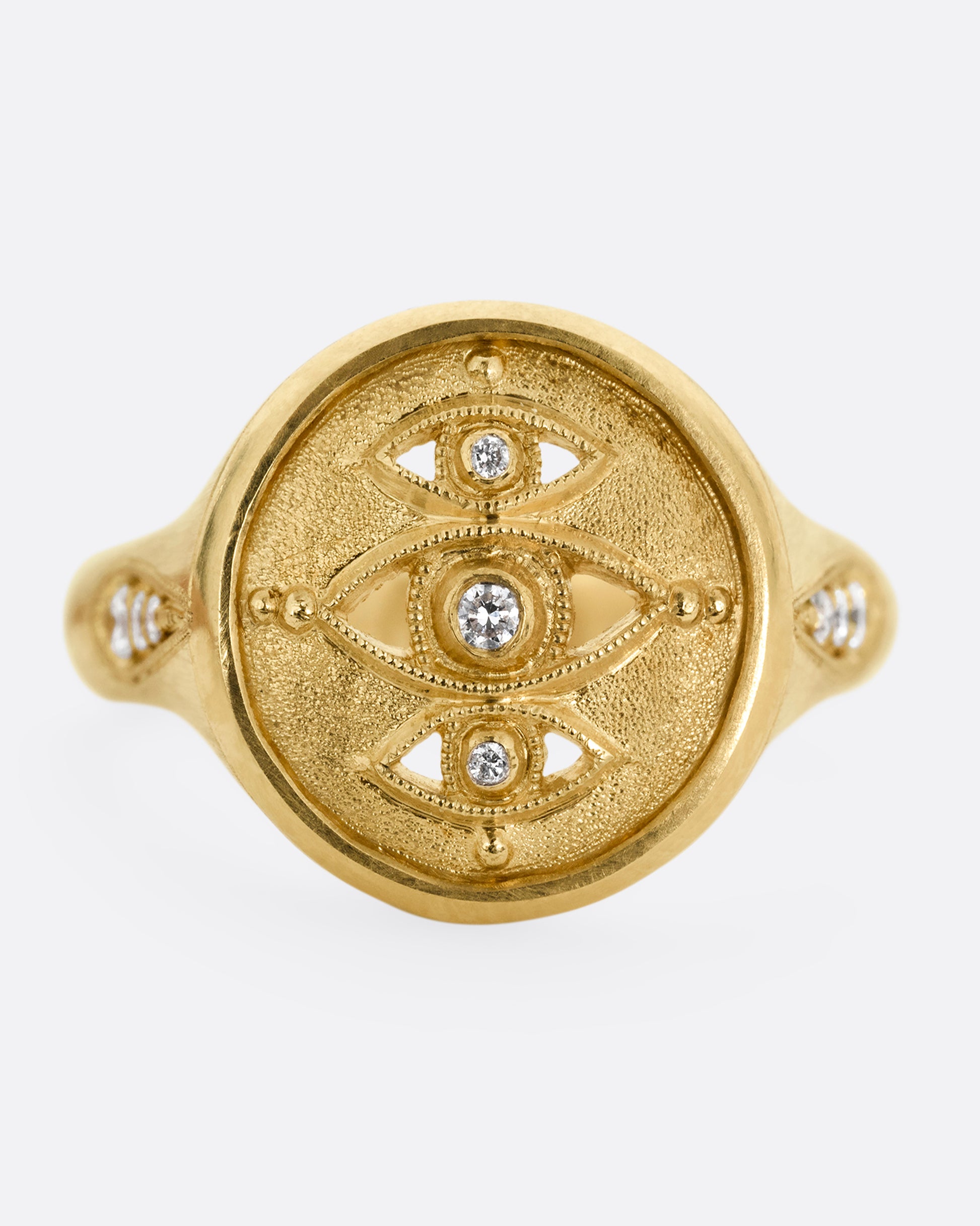 A yellow gold signet ring with three relief carved diamond eyes at the center. A trio of diamonds in teardrop shaped settings top each shoulder. Shown from the front.