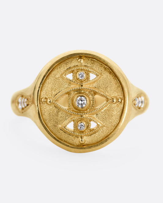 A yellow gold signet ring with three relief carved diamond eyes at the center. A trio of diamonds in teardrop shaped settings top each shoulder. Shown from the front.