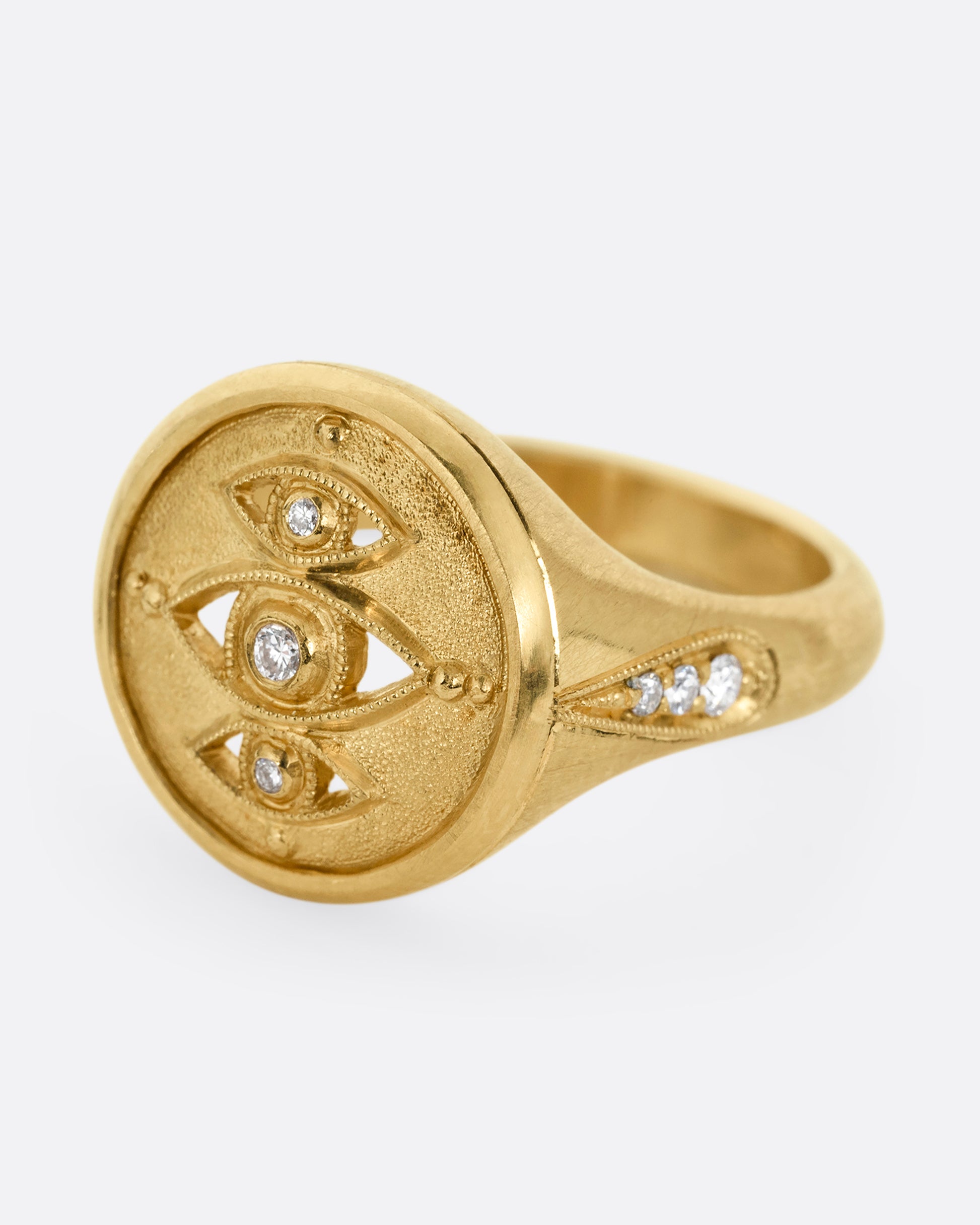 A yellow gold signet ring with three relief carved diamond eyes at the center. A trio of diamonds in teardrop shaped settings top each shoulder. Shown from the side.