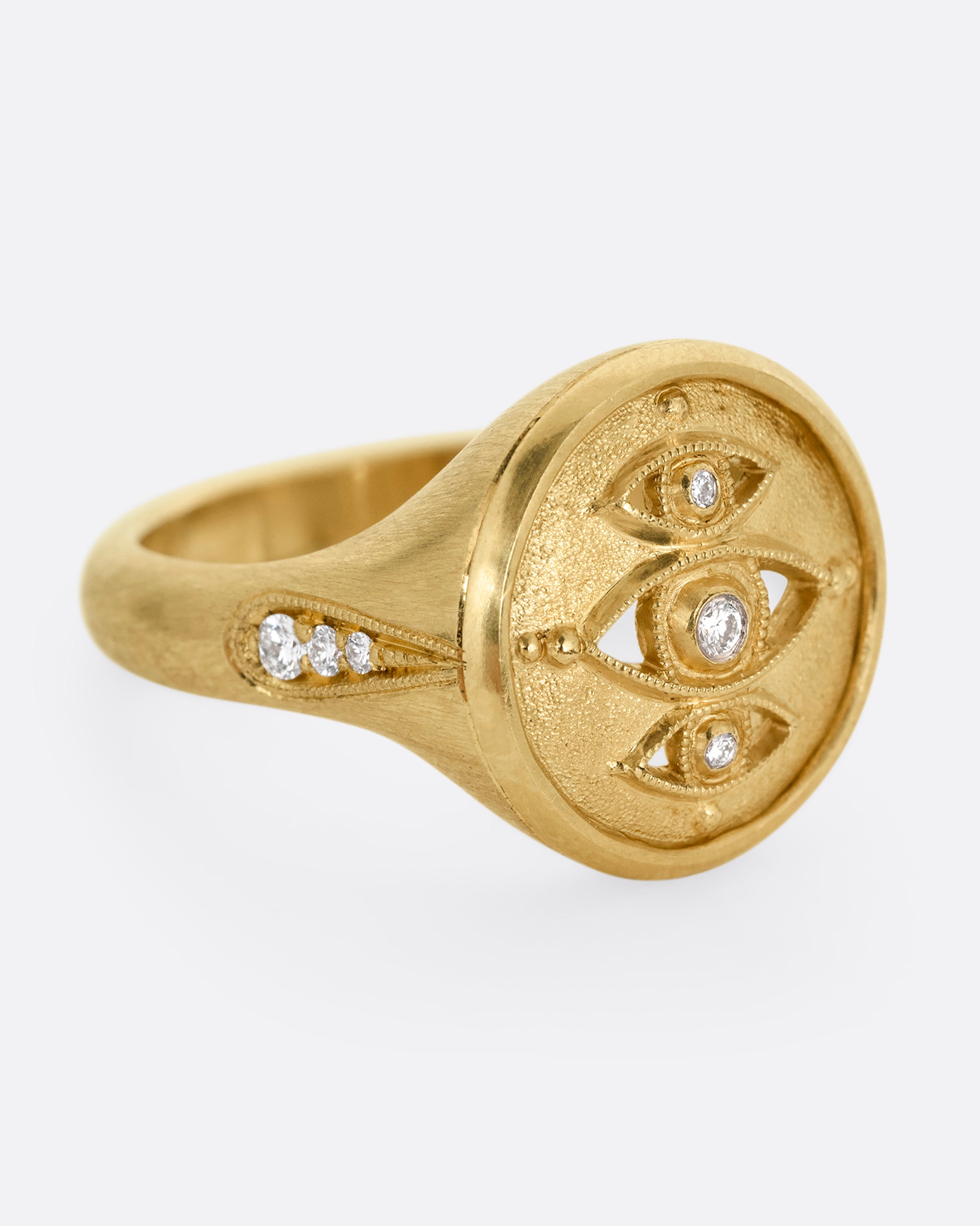 A yellow gold signet ring with three relief carved diamond eyes at the center. A trio of diamonds in teardrop shaped settings top each shoulder. Shown from the side.