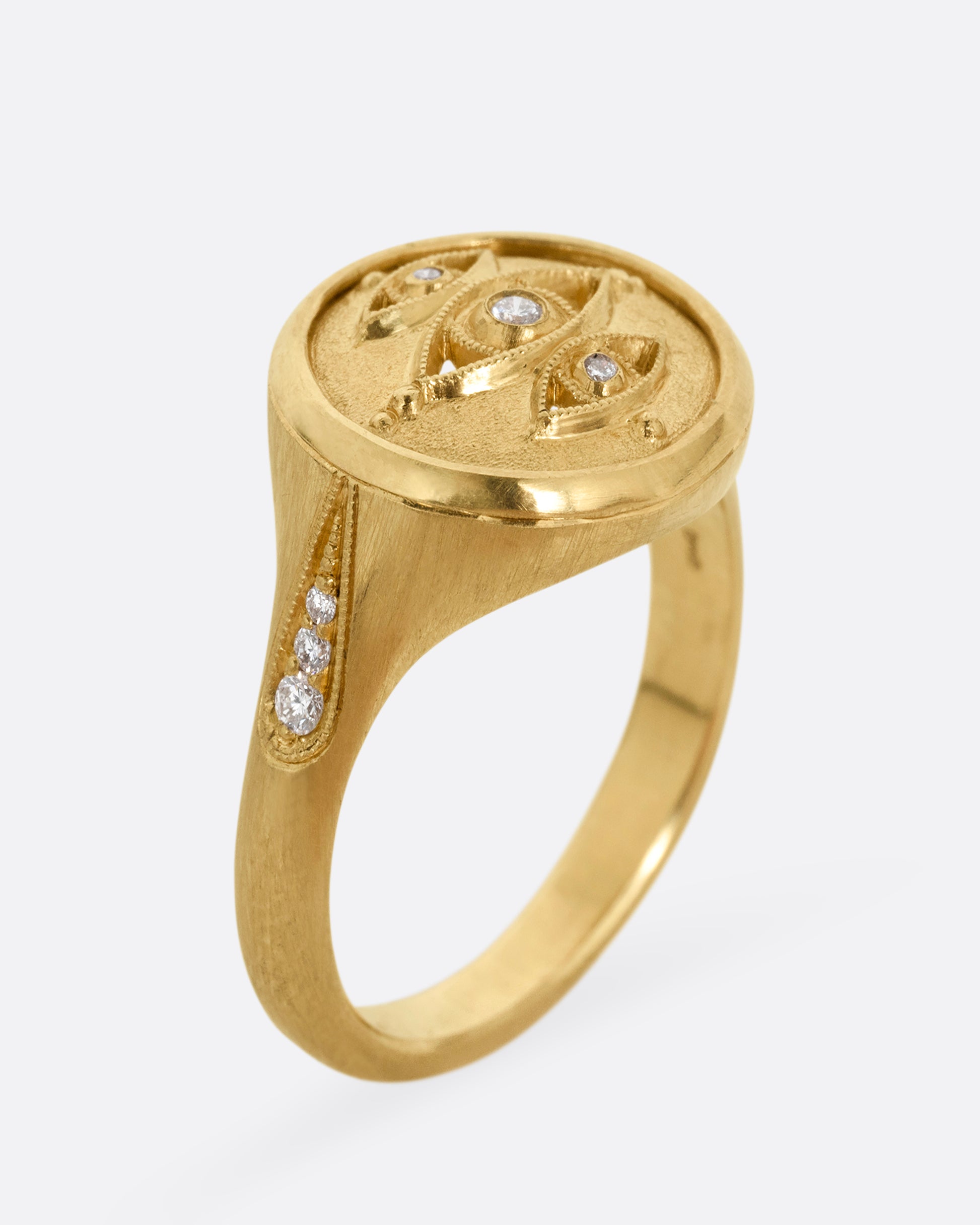 A yellow gold signet ring with three relief carved diamond eyes at the center. A trio of diamonds in teardrop shaped settings top each shoulder. Shown standing up from the side.