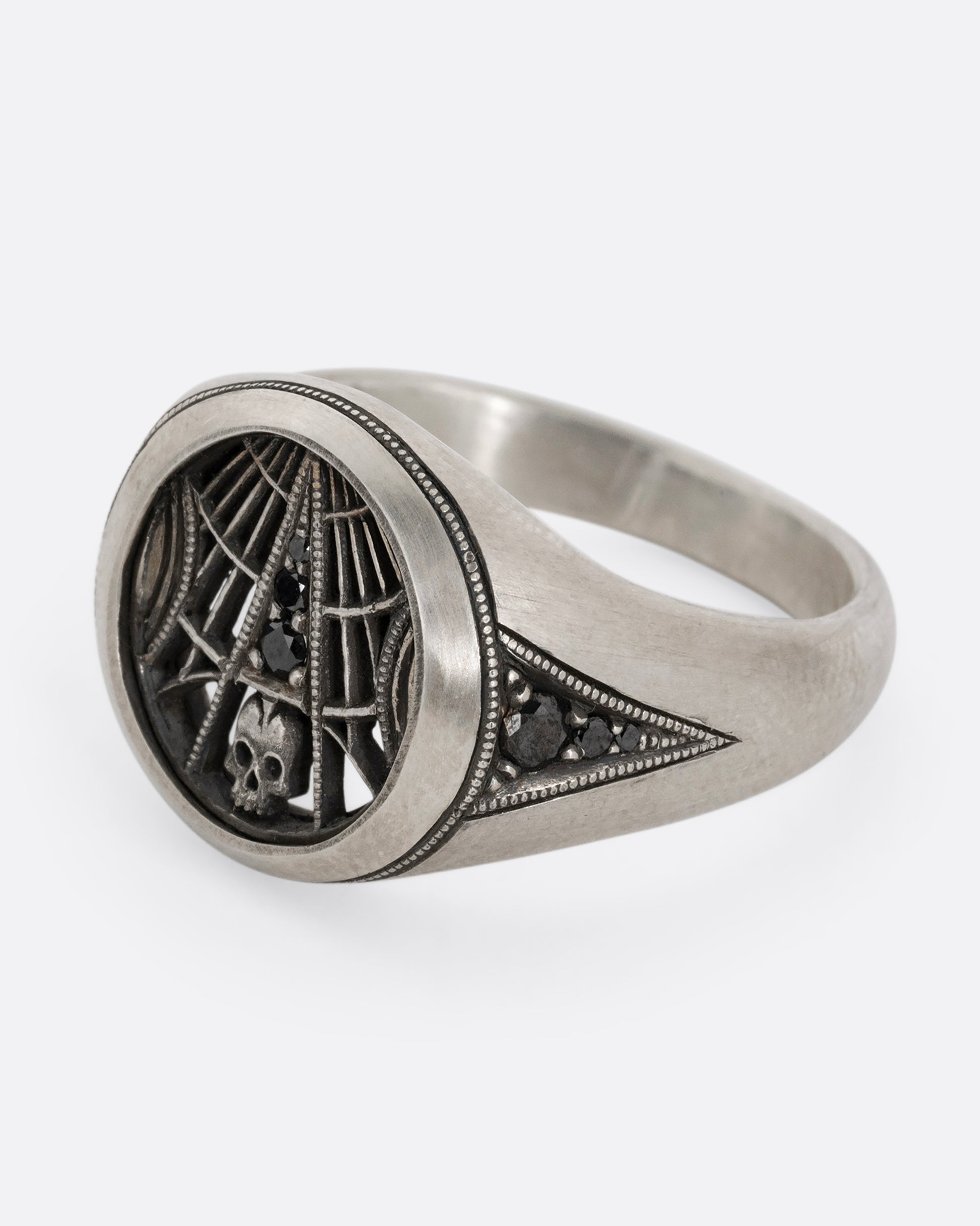 A silver signet ring with spider web details and black diamonds stacked atop a tiny skull. Shown from the side.
