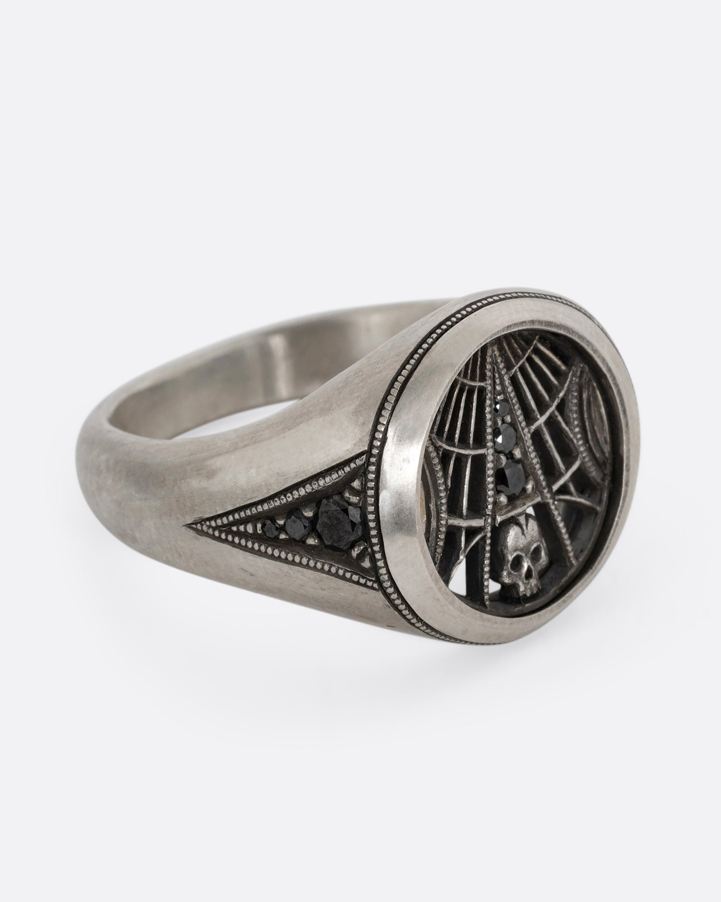 A silver signet ring with spider web details and black diamonds stacked atop a tiny skull. Shown from the side.