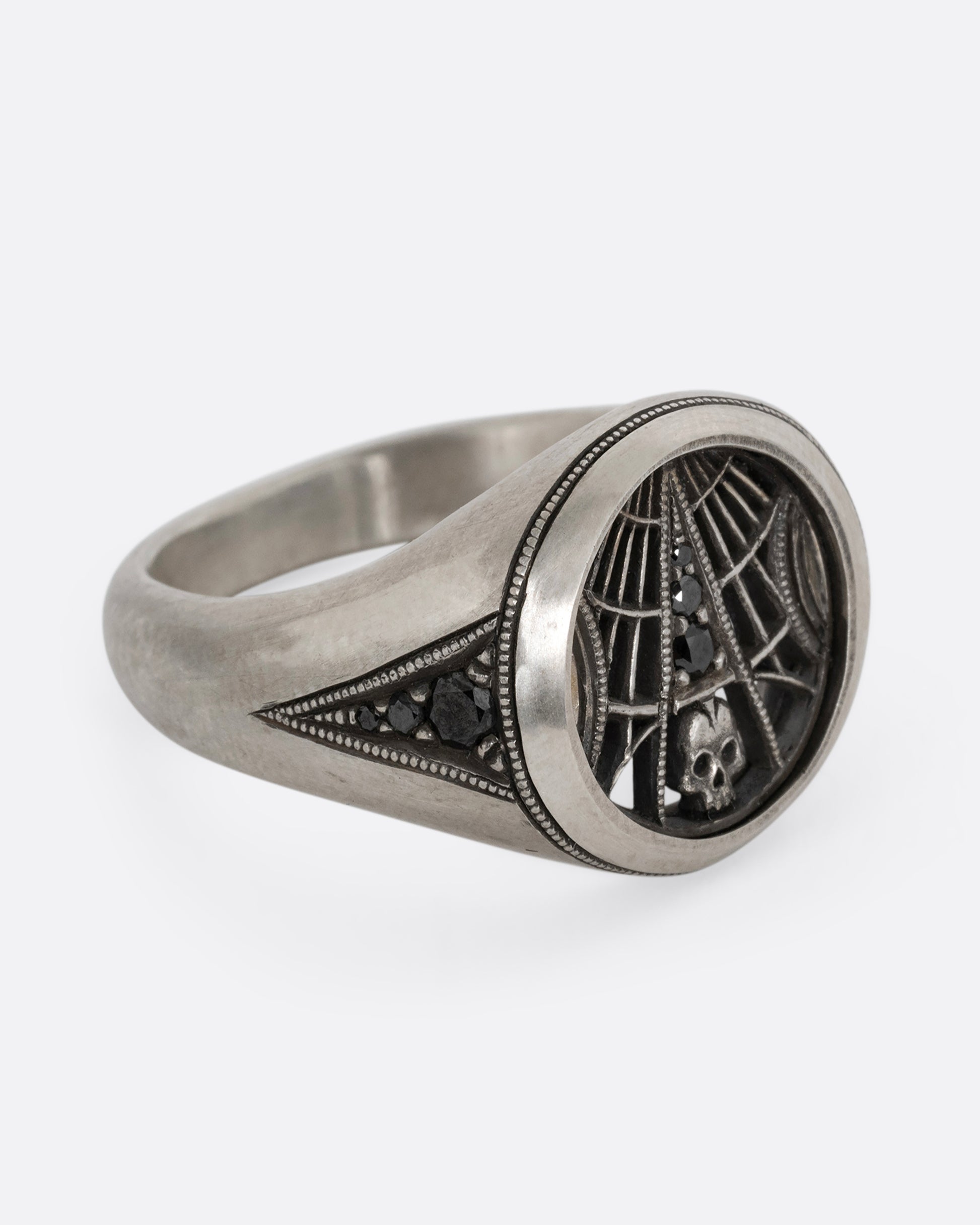 A silver signet ring with spider web details and black diamonds stacked atop a tiny skull. Shown from the side.