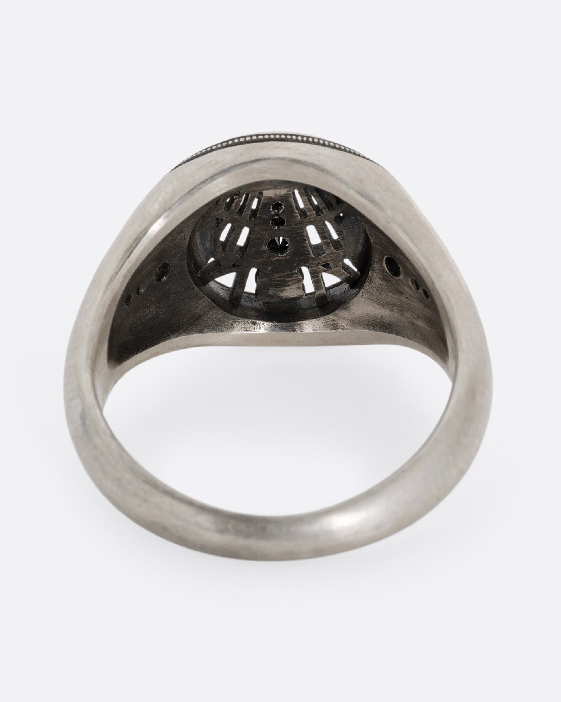 A silver signet ring with spider web details and black diamonds stacked atop a tiny skull. Shown from the back.