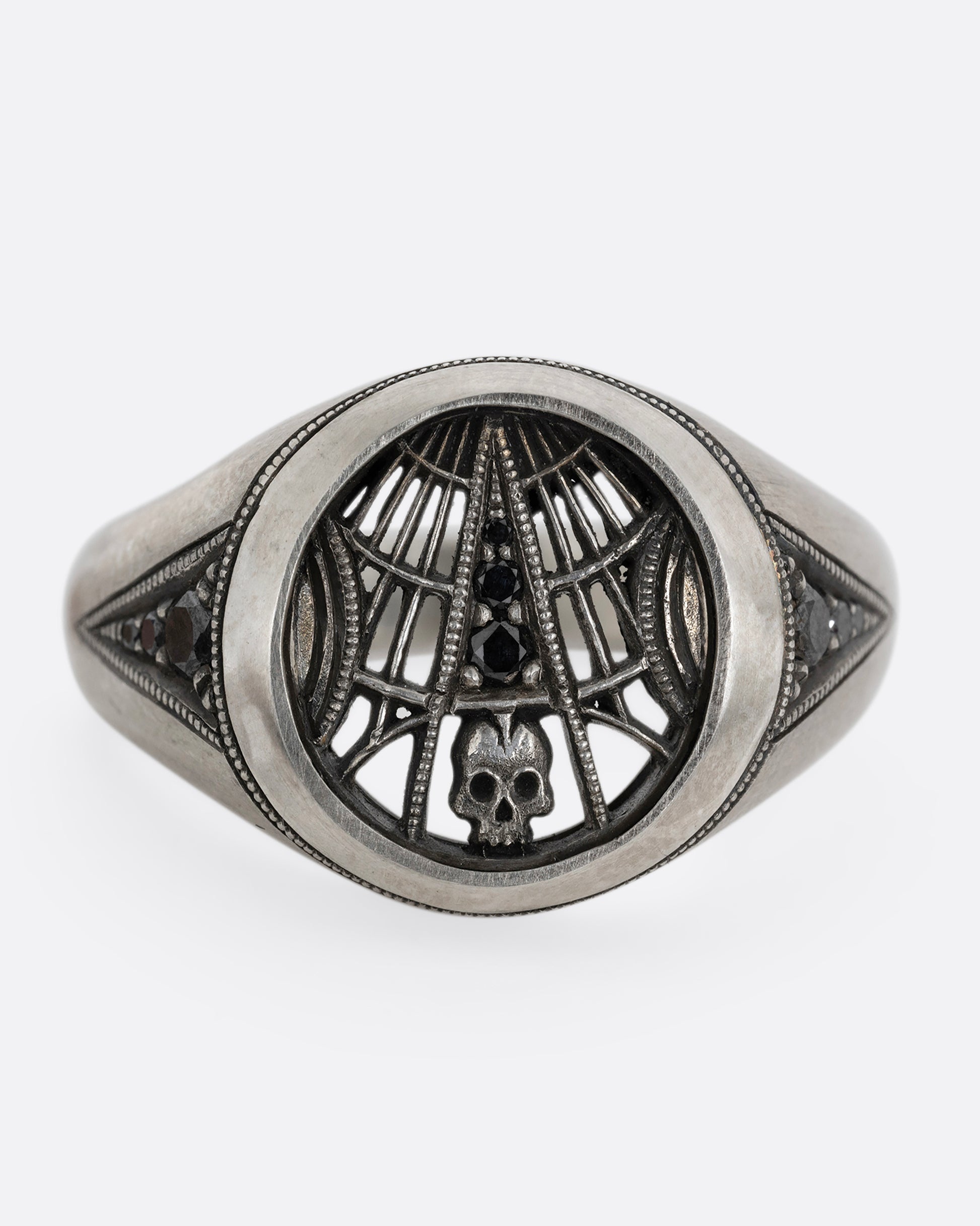 A silver signet ring with spider web details and black diamonds stacked atop a tiny skull. Shown from the front.