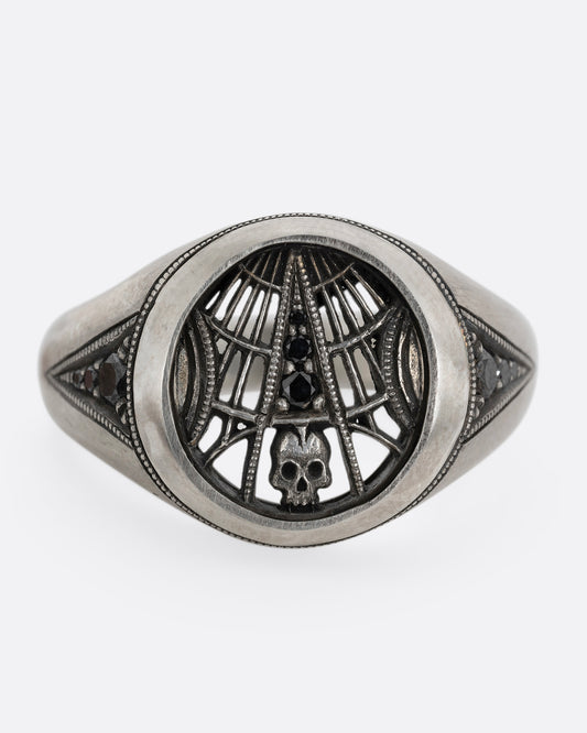 A silver signet ring with spider web details and black diamonds stacked atop a tiny skull. Shown from the front.
