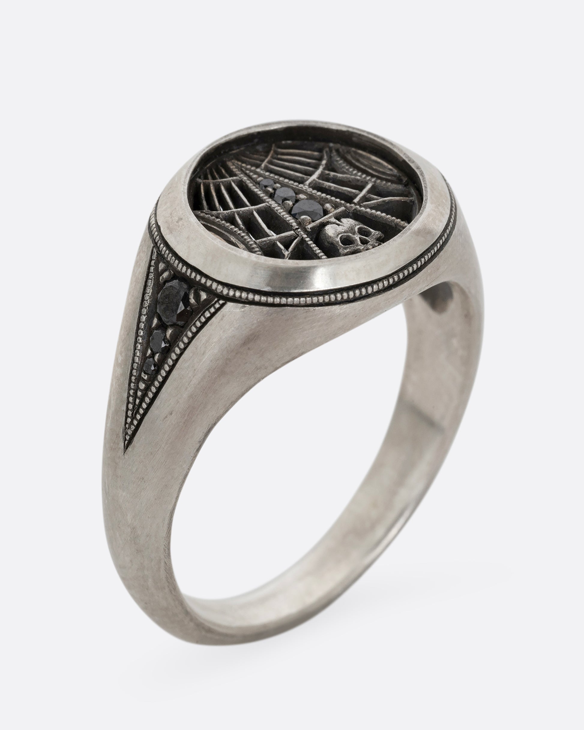 A silver signet ring with spider web details and black diamonds stacked atop a tiny skull. Shown standing up diagonally from the front.