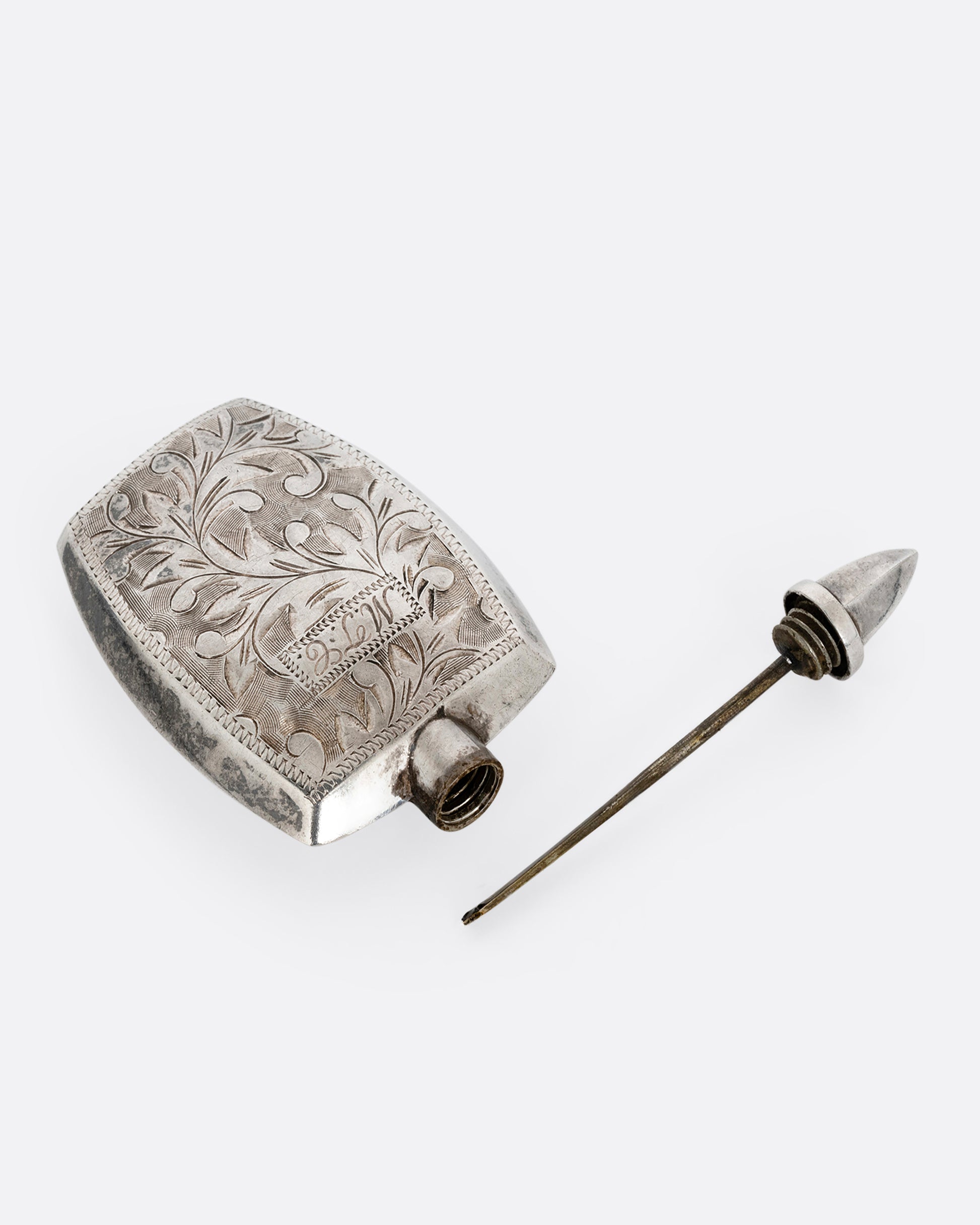 A silver perfume bottle with botanical etchings, a twist- off wand, and the engraved initials M.T.C. Shown open from the top.