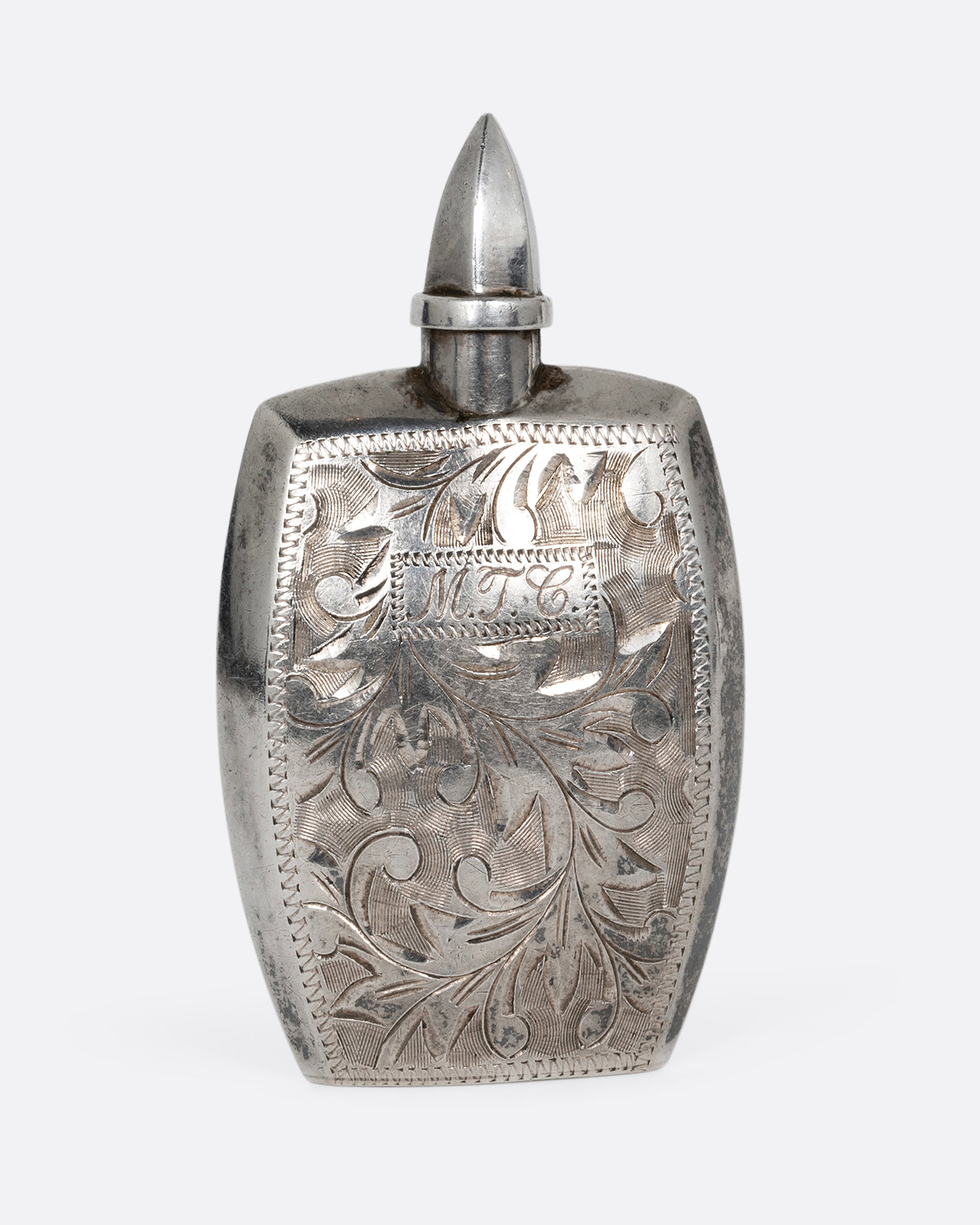 A silver perfume bottle with botanical etchings, a twist- off wand, and the engraved initials M.T.C. Shown from the front.