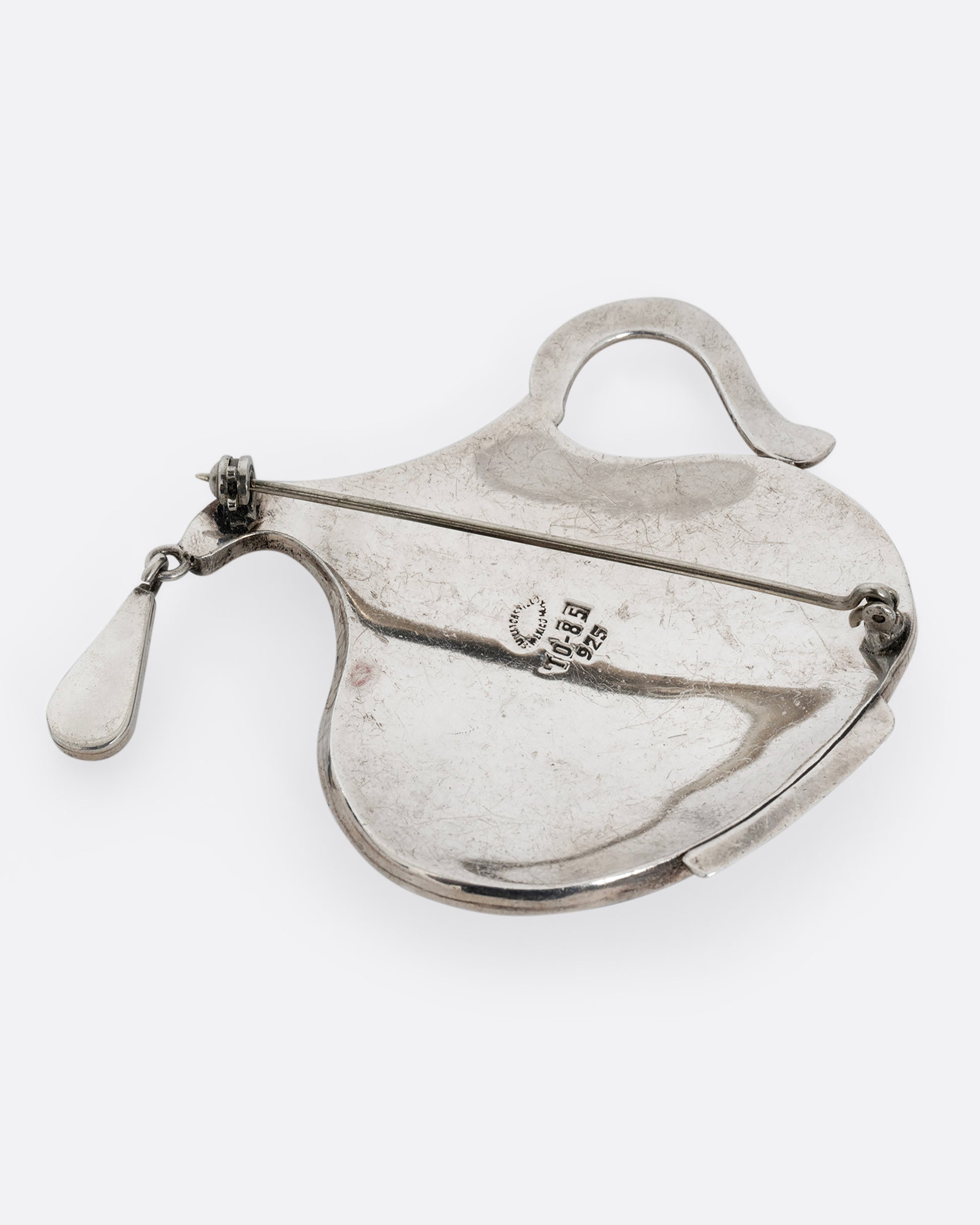 A sterling silver pin in the shape of a pitcher filled with water made of lapis and a dangling drop. Shown laying flat.
