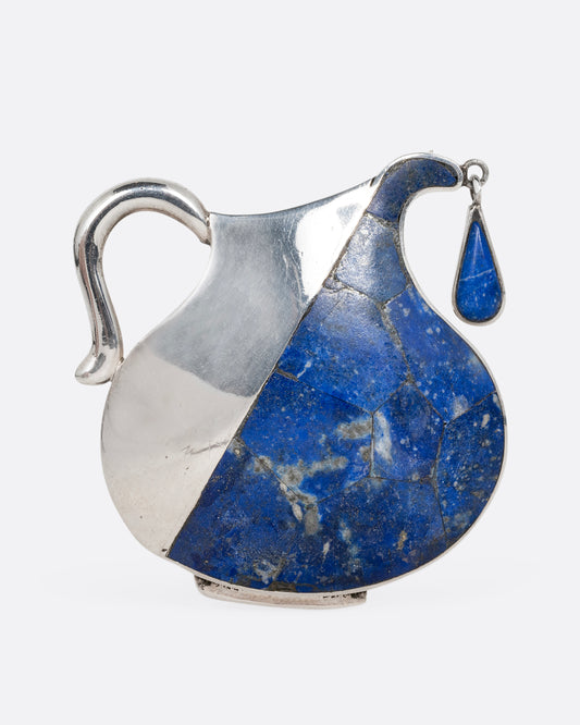 A sterling silver pin in the shape of a pitcher filled with water made of lapis and a dangling drop. Shown from the front.
