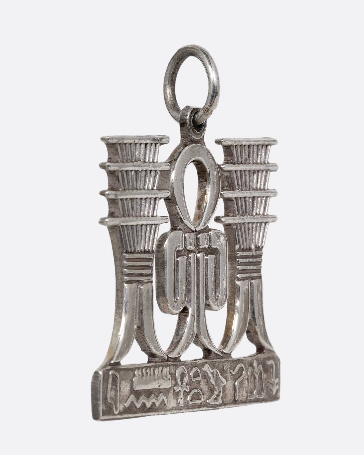 A silver pendant with Egyptian symbols of Sa and Djed with hieroglyphs along the bottom. Shown from the side.