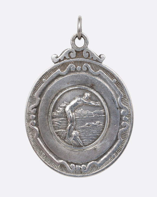 A silver medallion with a swimmer preparing to dive at the center, surrounded by filigree. Shown from the front.