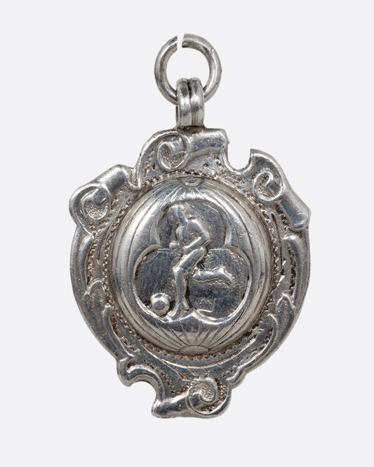 A silver pendant with a soccer player framed by intricate scrollwork. Shown from the front.