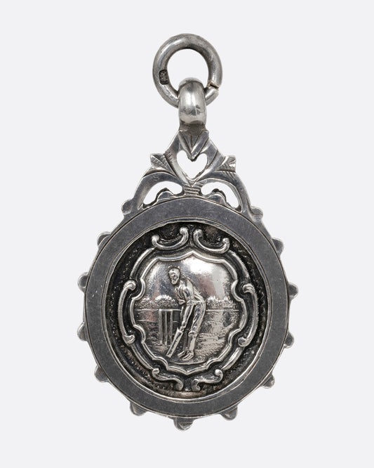 A round silver pendant with a cricket batter mid-swing surrounded by filigree detailing. Shown from the front.