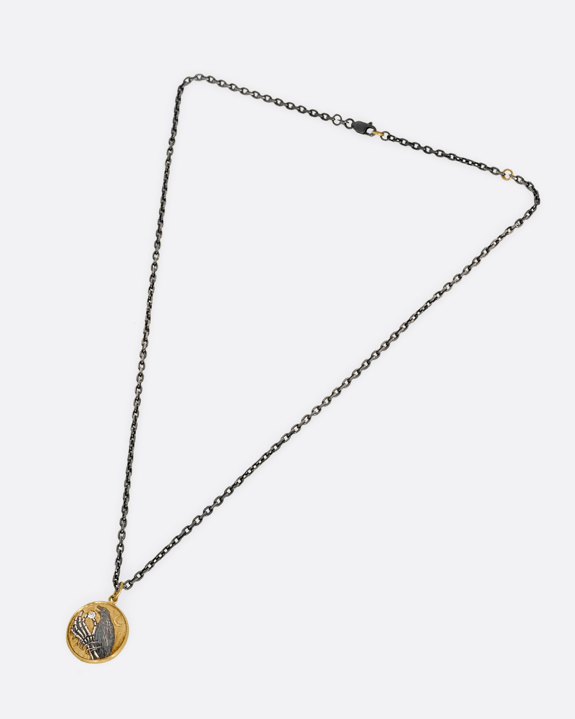 A yellow gold and silver pendant necklace with a skeleton hand holding a diamond and a crow. Shown laying flat from the front.