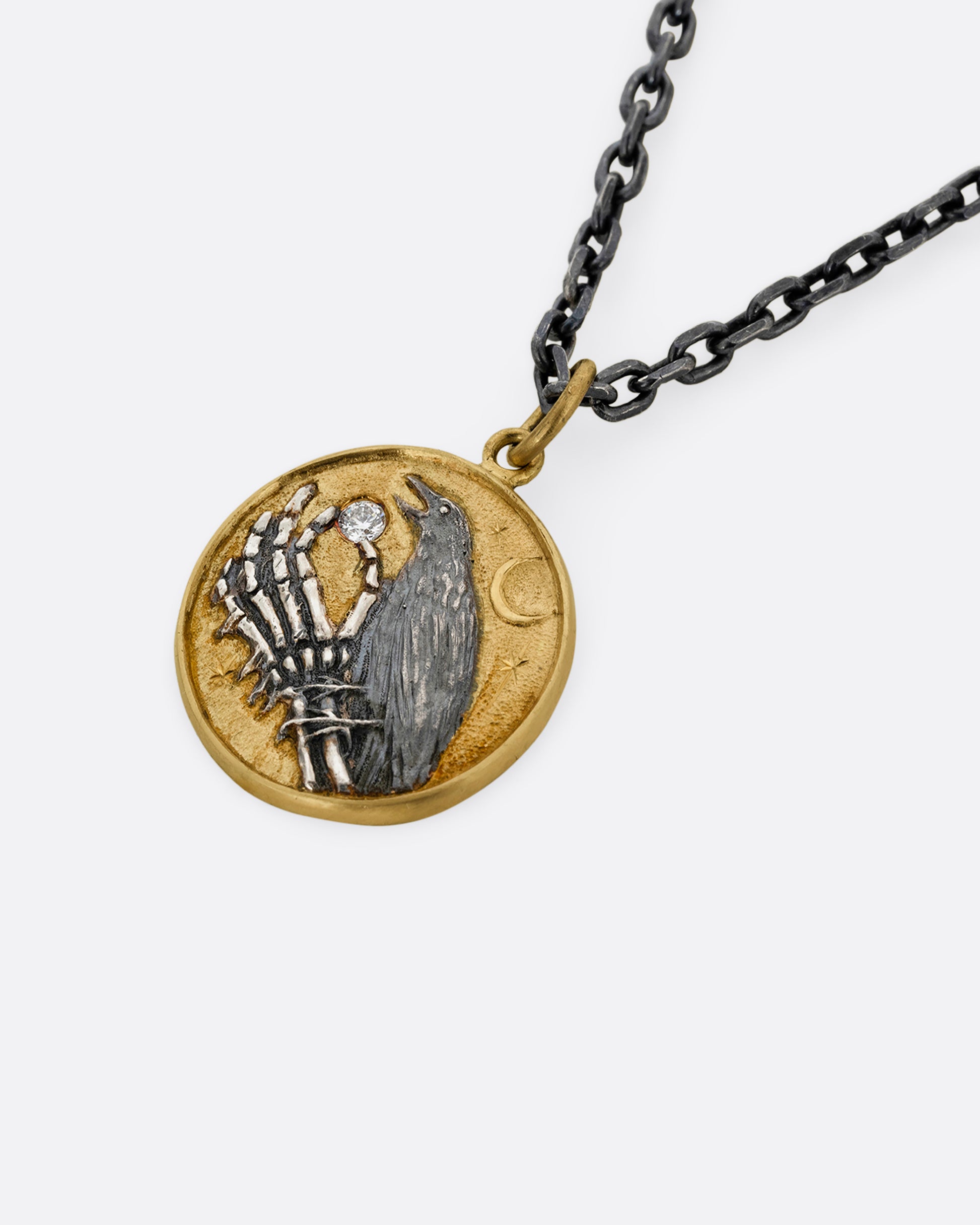 A yellow gold and silver pendant necklace with a skeleton hand holding a diamond and a crow. Shown close up from the front.
