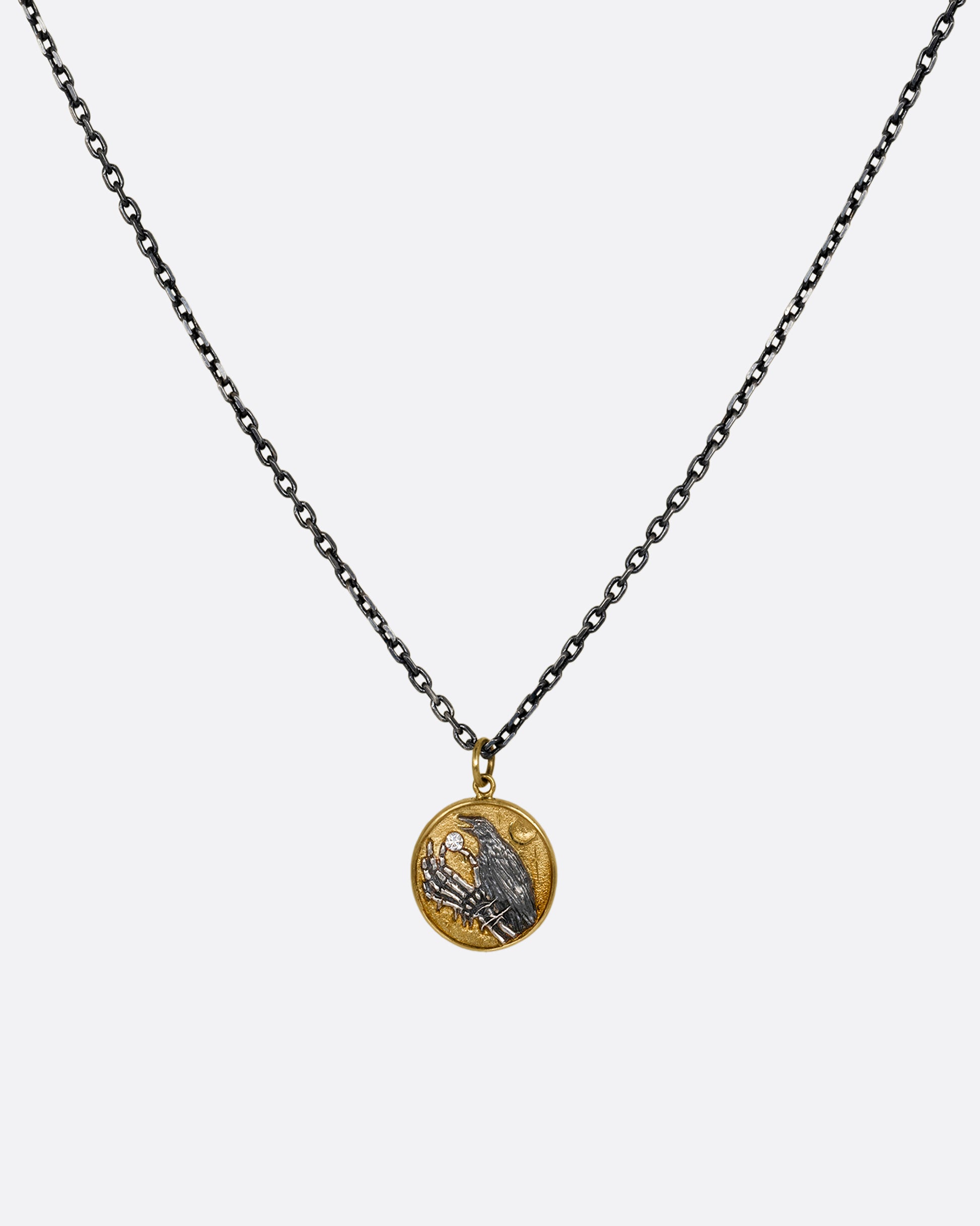 A yellow gold and silver pendant necklace with a skeleton hand holding a diamond and a crow. Shown from the front.