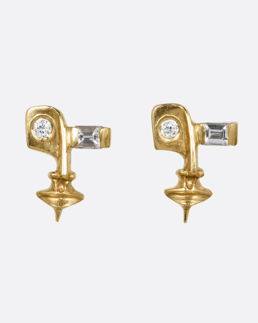 A pair of yellow gold stud earrings with cubist inspired faces with one flush set diamond eye and one baguette diamond eye. Shown from the front.