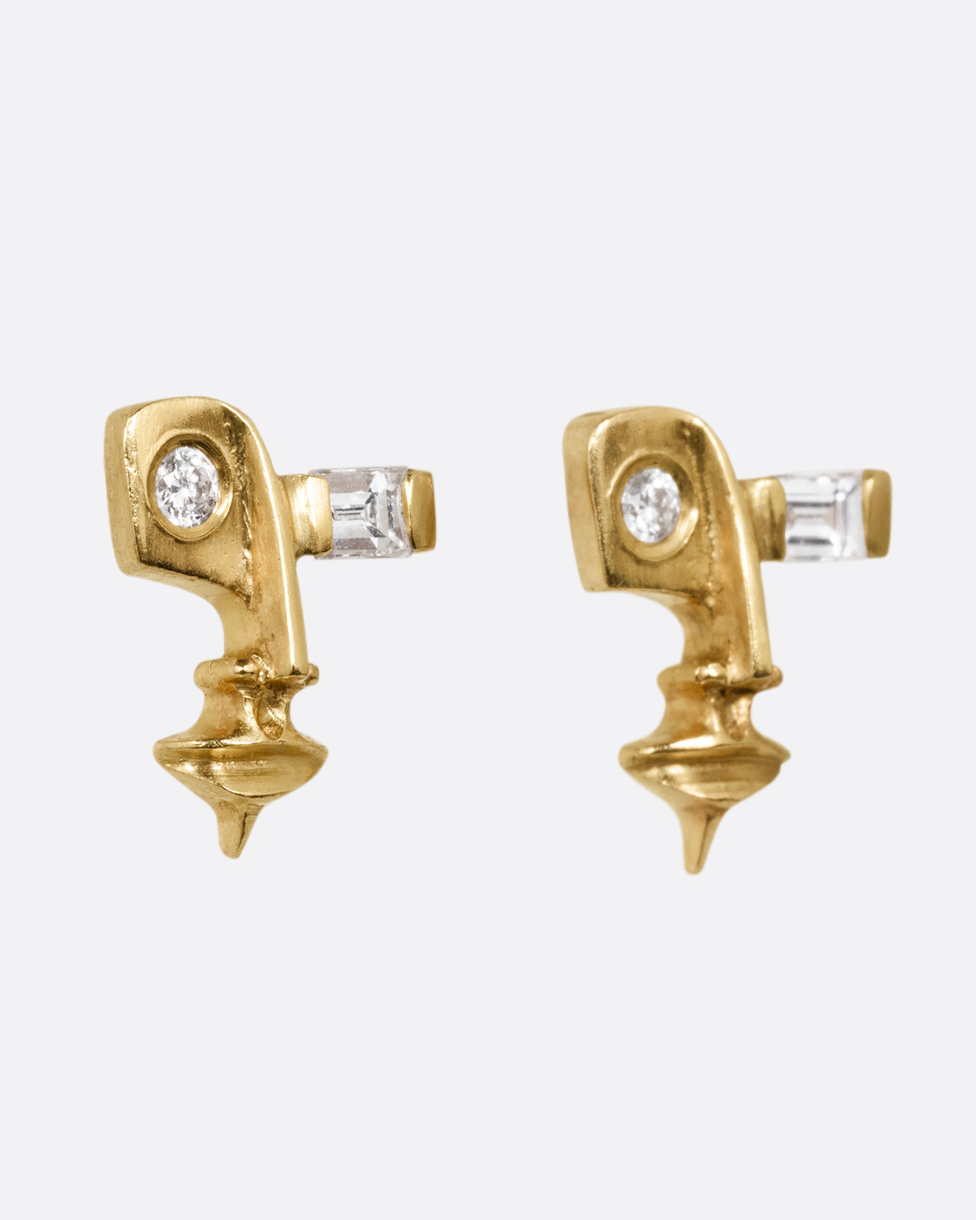 A pair of yellow gold stud earrings with cubist inspired faces with one flush set diamond eye and one baguette diamond eye. Shown from the side.