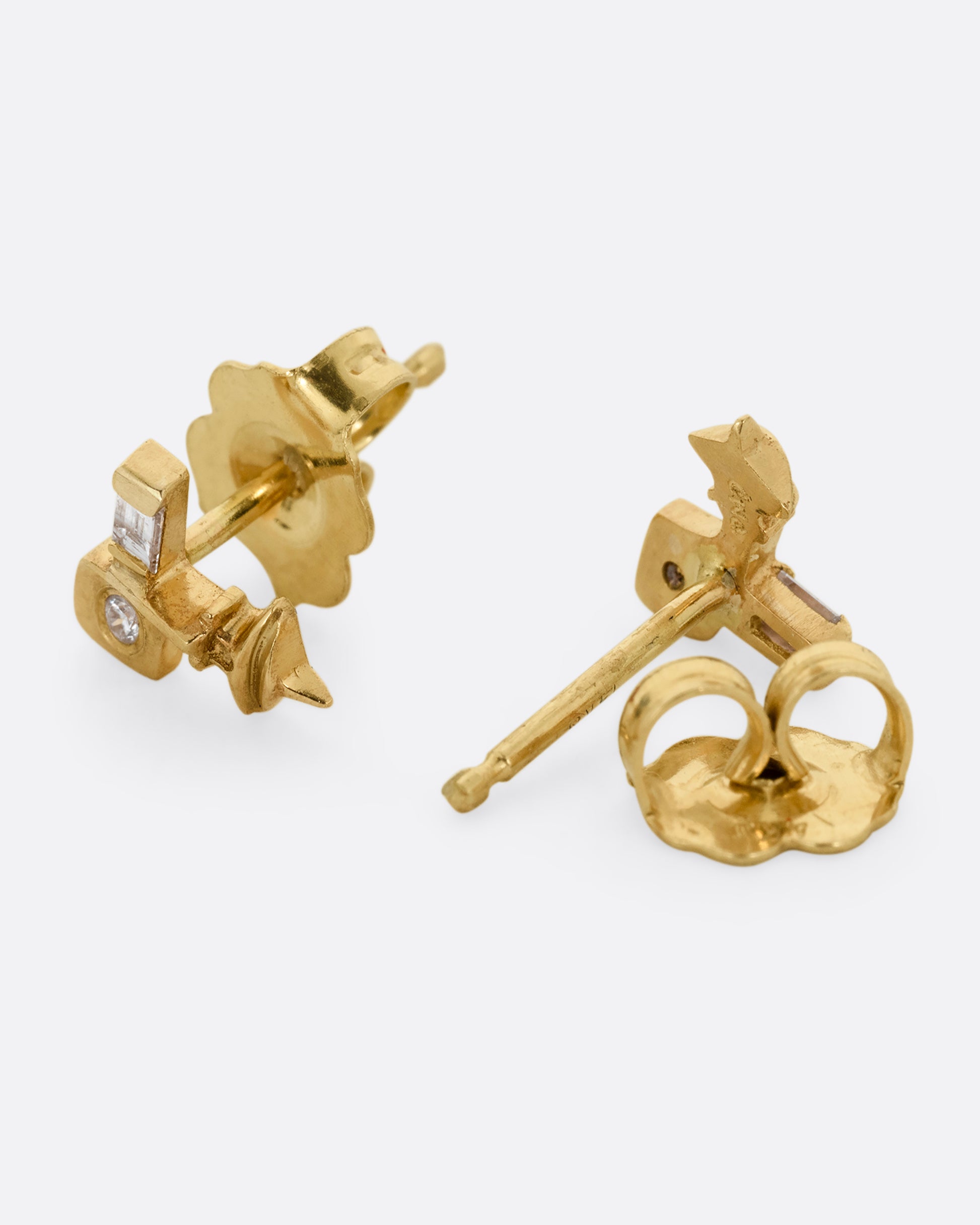 A pair of yellow gold stud earrings with cubist inspired faces with one flush set diamond eye and one baguette diamond eye. One earring is shown on its side from the front. The other is shown upside down from the back with an open stud back.