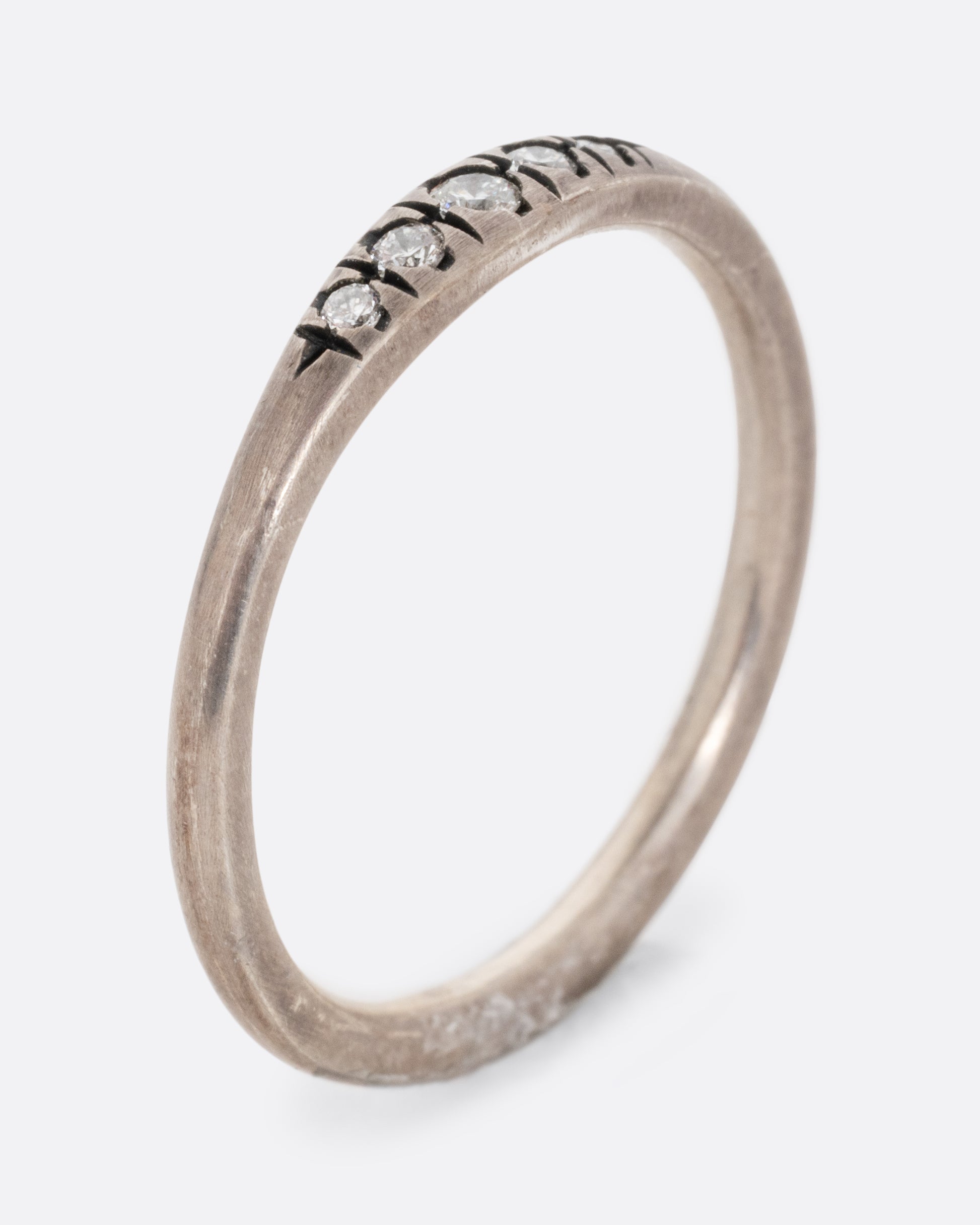 A silver ring with white diamonds and darkened engravings. Shown standing up from the side.