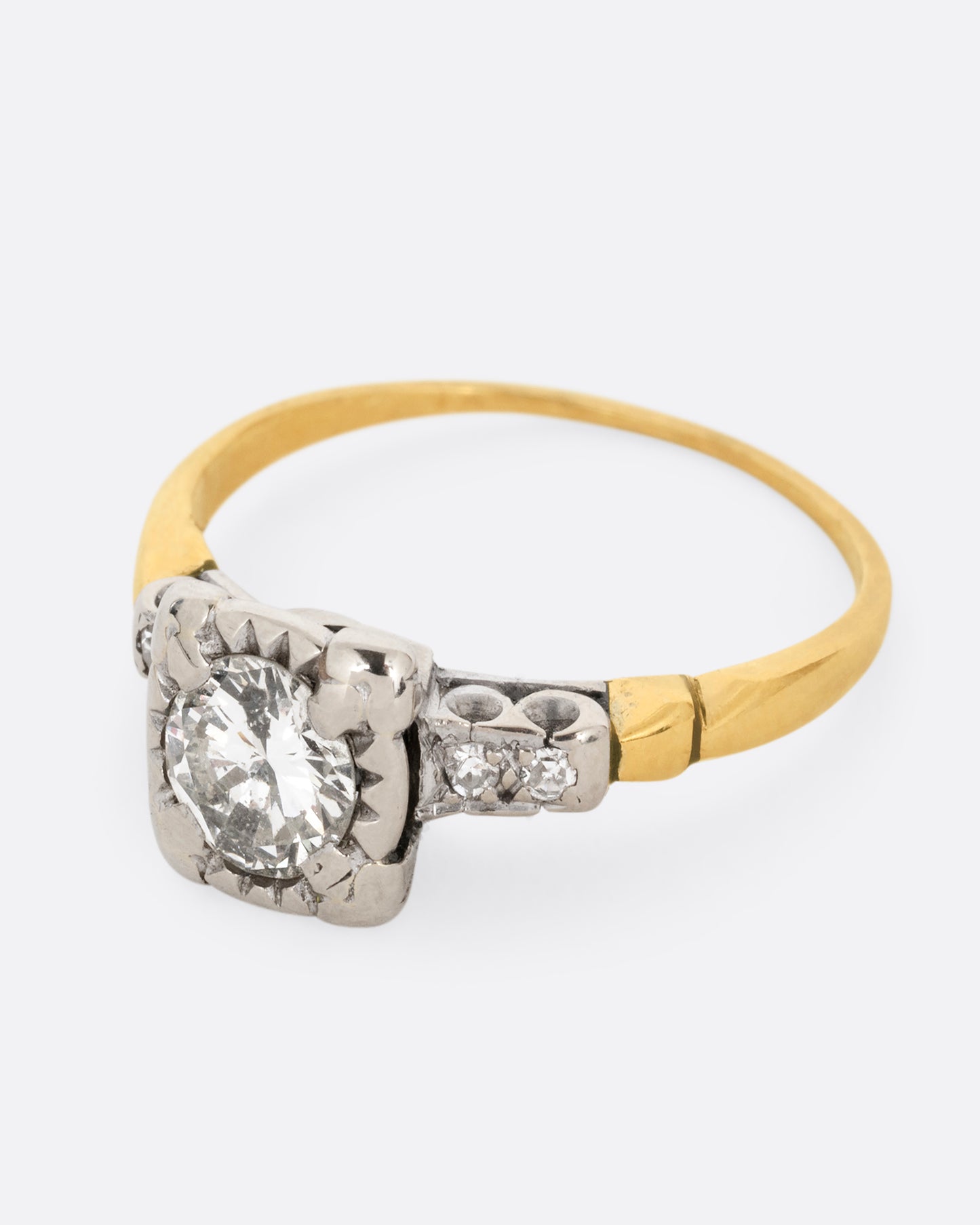 A yellow gold ring with a white gold setting and a round central diamond with four bead set diamonds on the shoulders. Shown from the side.
