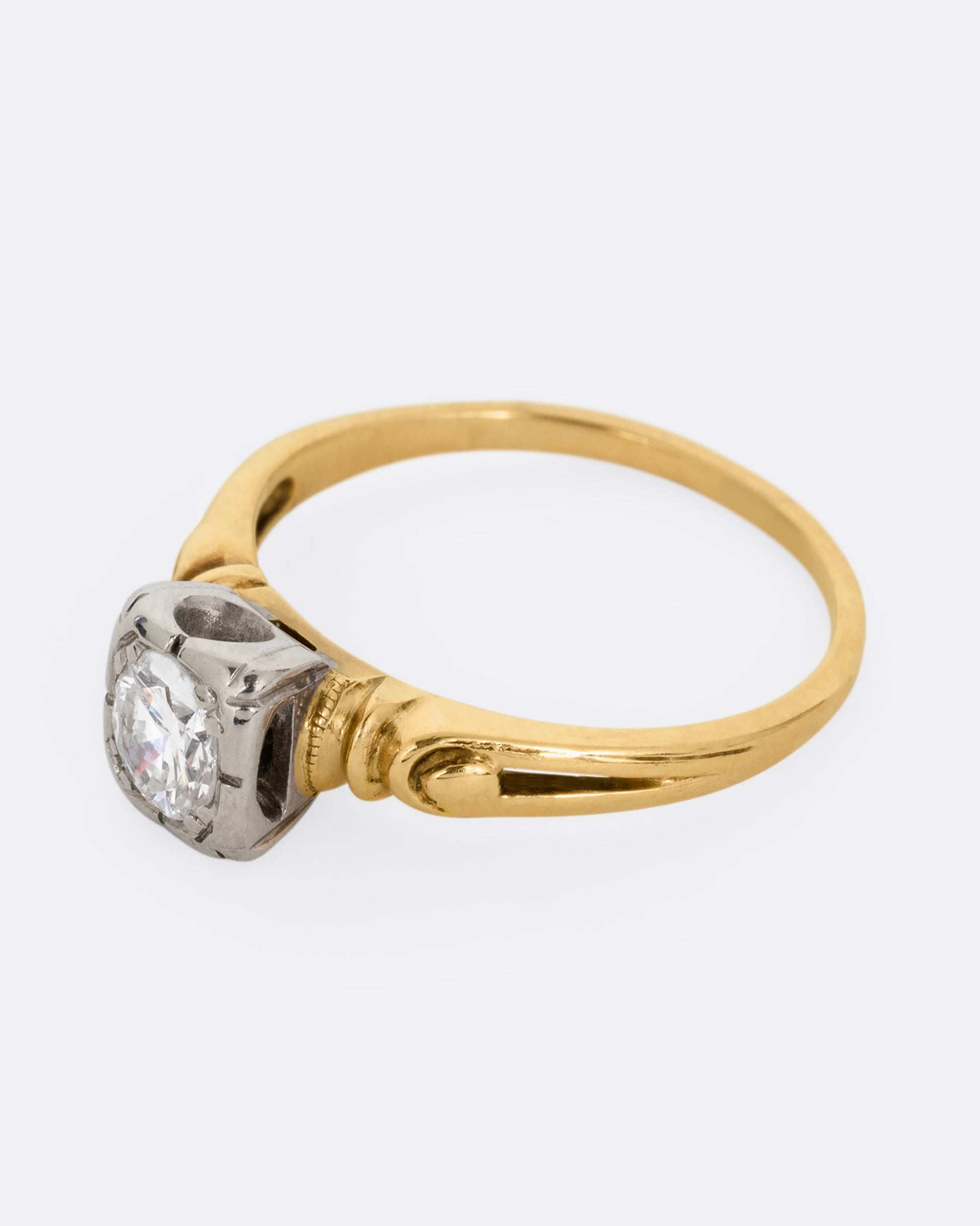 A yellow gold ring with intricately detailed shoulders and a white gold set center diamond. Shown from the side.
