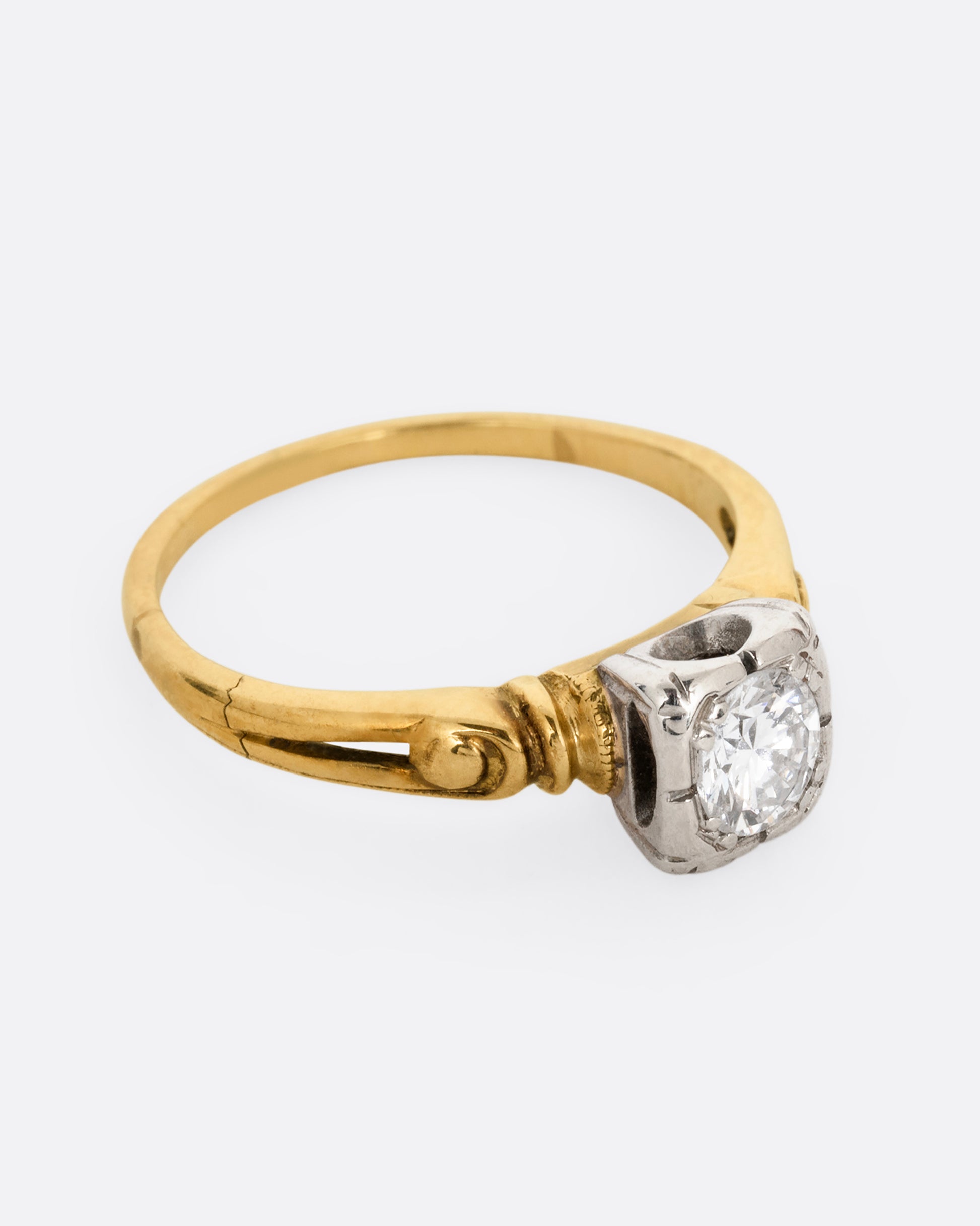 A yellow gold ring with intricately detailed shoulders and a white gold set center diamond. Shown from the side.