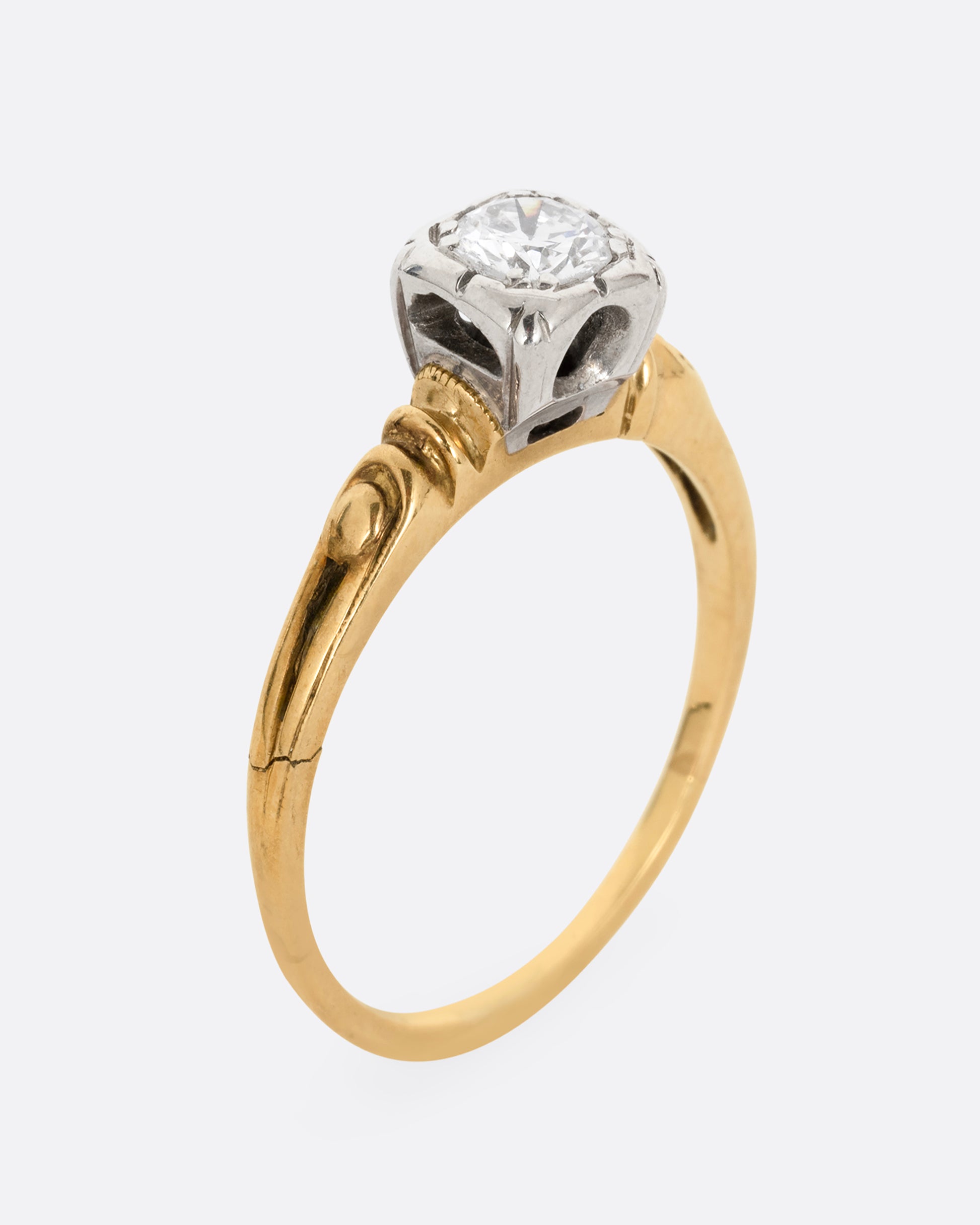 A yellow gold ring with intricately detailed shoulders and a white gold set center diamond. Shown standing up from the side.