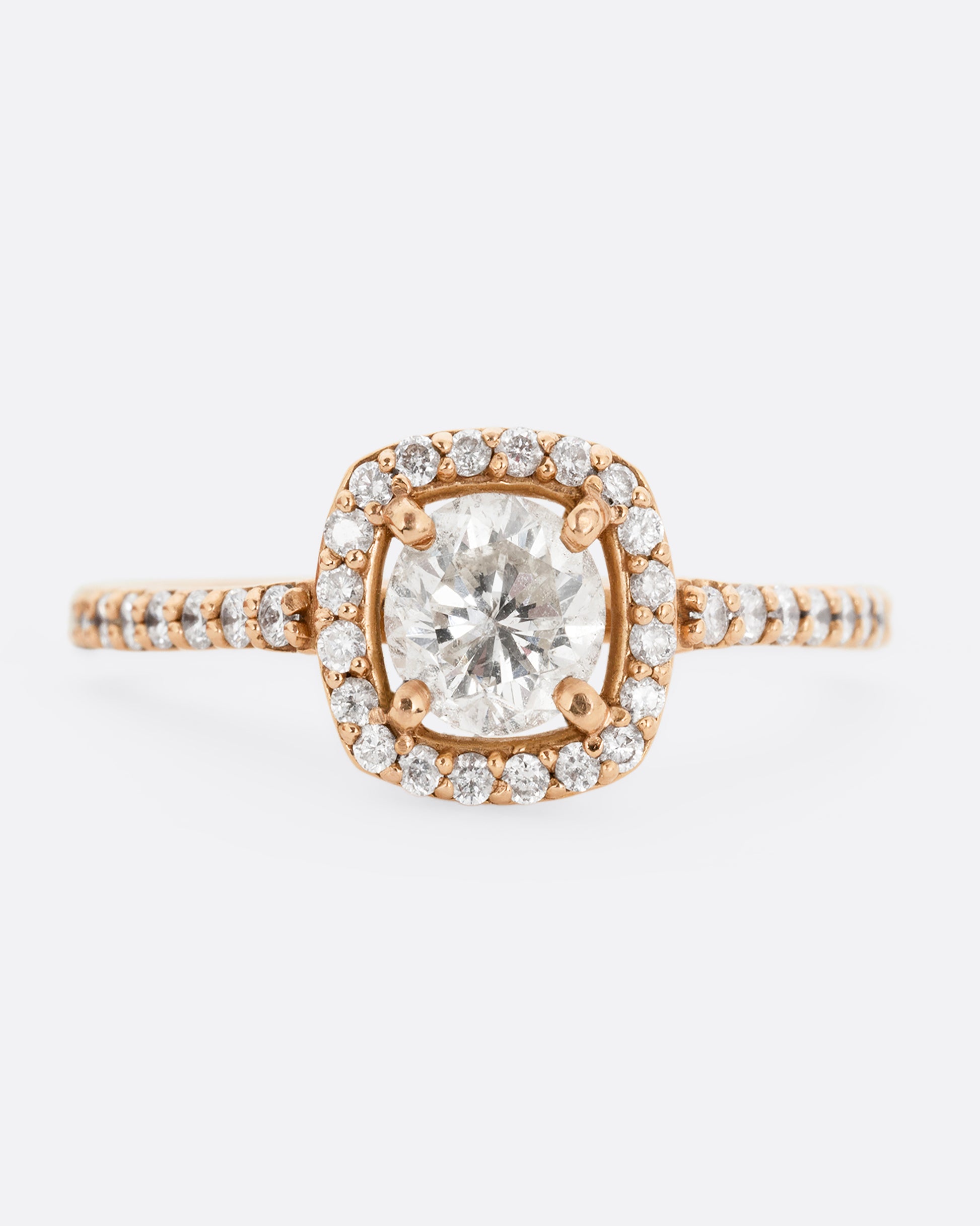 A rose gold ring with a  prong set diamond surrounded by pave diamonds. The pave also wraps three quarters around the band. Shown from the front.