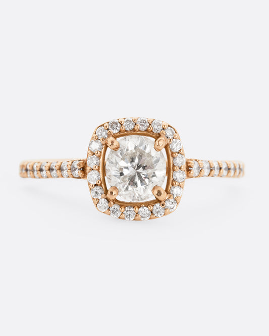 A rose gold ring with a  prong set diamond surrounded by pave diamonds. The pave also wraps three quarters around the band. Shown from the front.