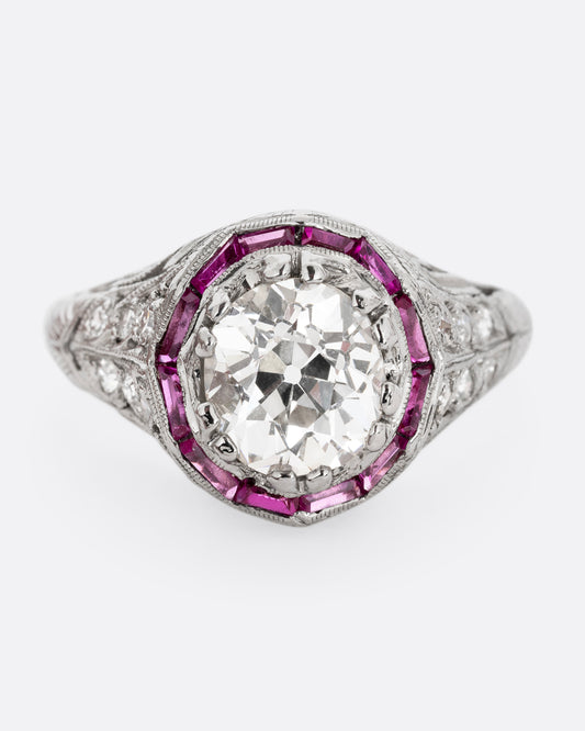 A platinum ring with an old European cut diamond surrounded by rubies and white diamond accents. Shown from the front.