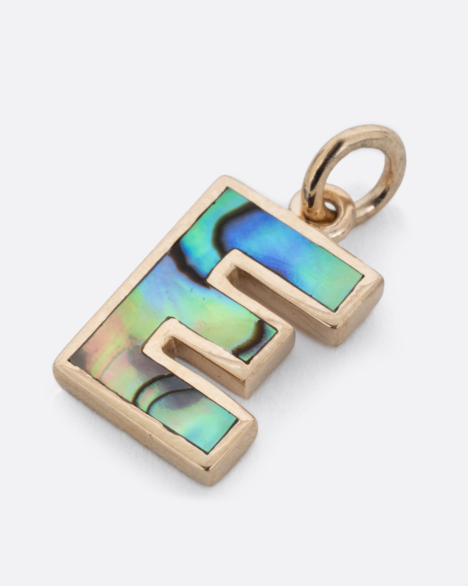 An abalone E charm set in yellow gold with a yellow gold jump ring and hoop. Shown lying flat at a diagonal from the front.