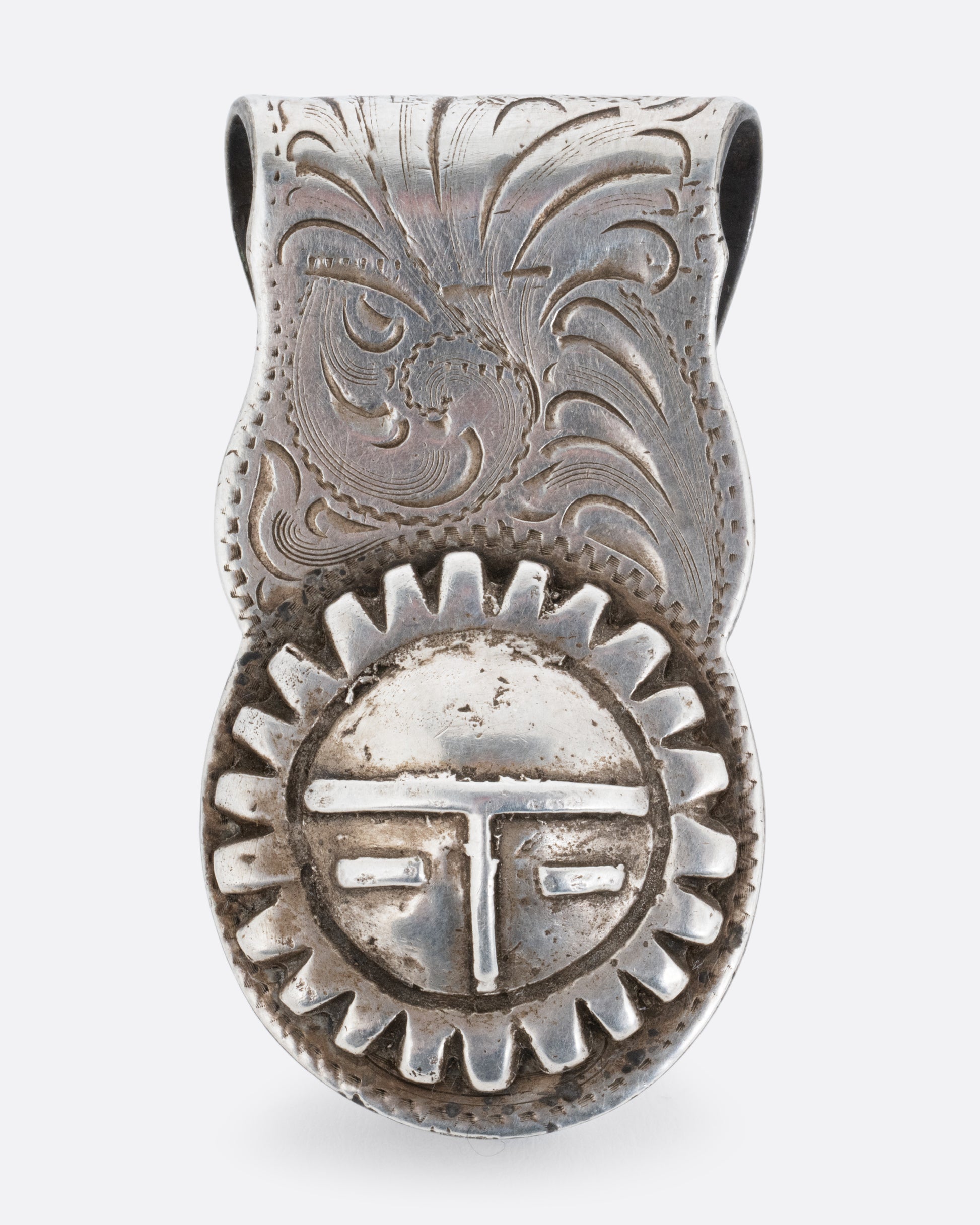 A silver money clip with a swirling engraved pattern and the Hopi sun spirit and creator, Tawa, on the end of the front. Shown from the front.
