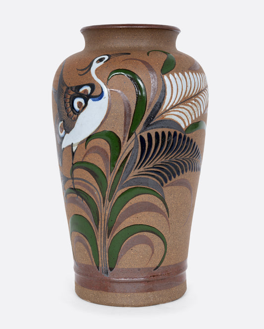 A tall ceramic vase with a hand painted bird among flourishes of ferns. Shown from the front.